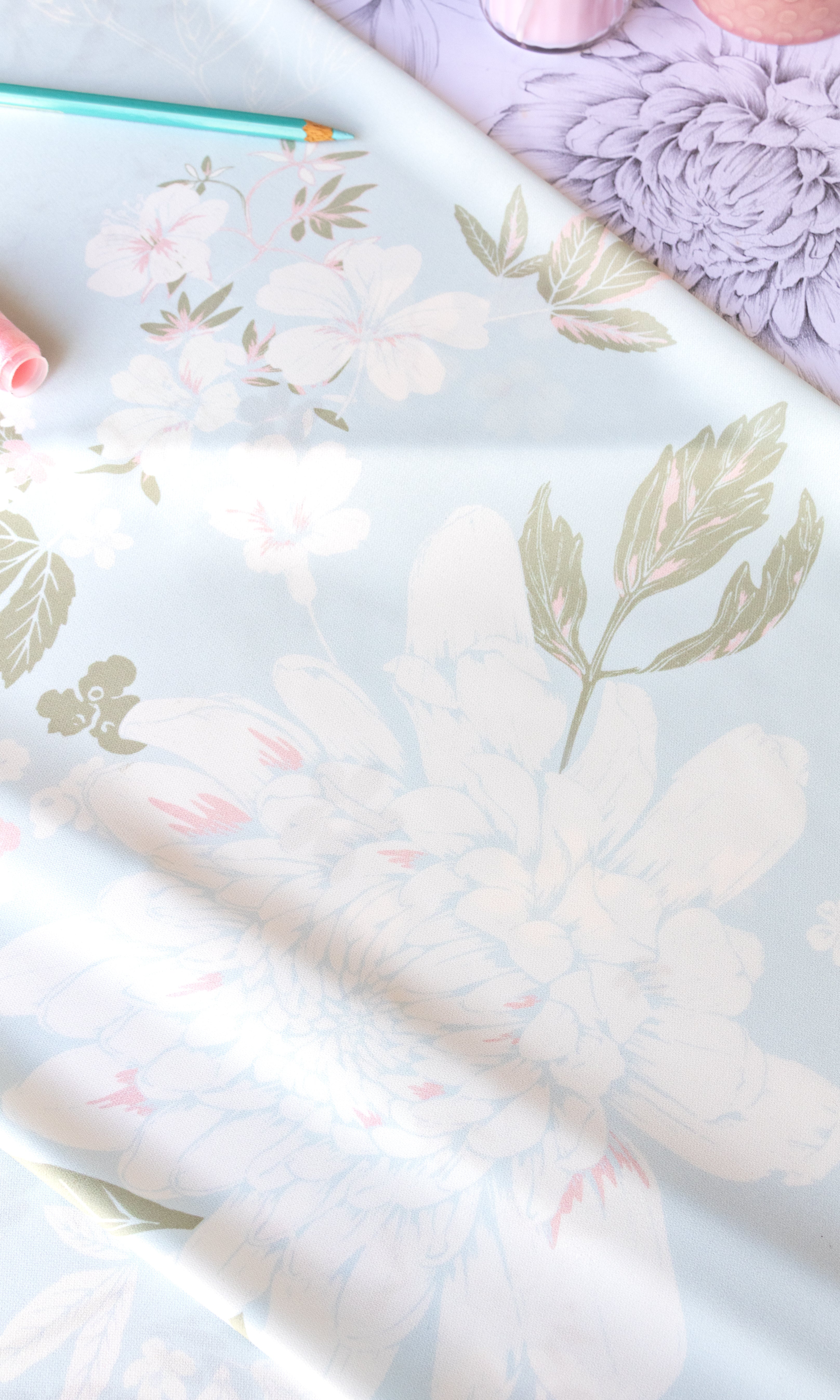A close-up of a floral fabric or wallpaper sample with large pale pink and white flowers, muted green leaves, and a soft lavender background. A teal pencil and pink decorative items are partially visible at the top edge.