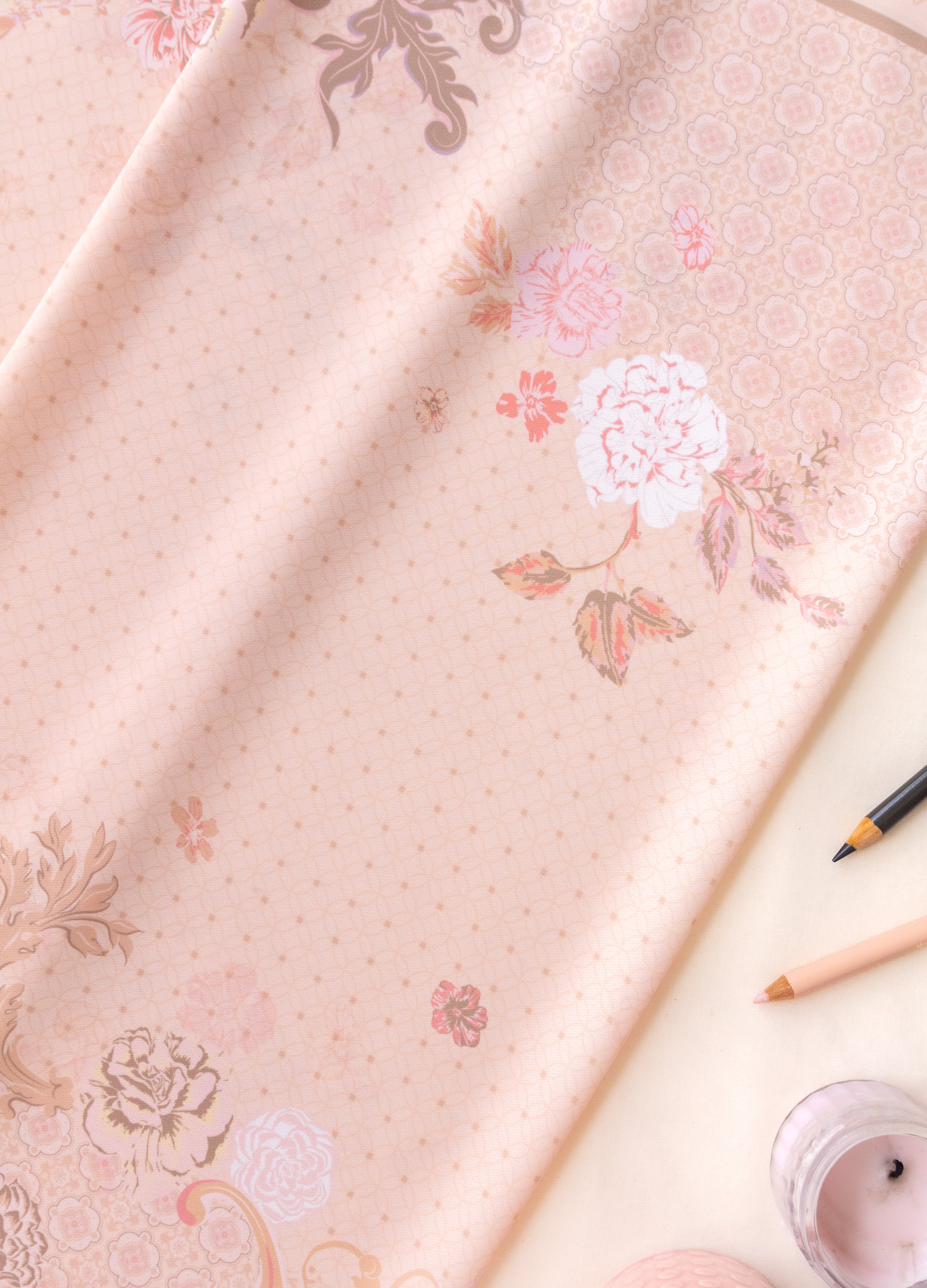 A light pink fabric with a delicate floral and scroll pattern, styled in a flat-lay composition with a pencil, a candle, and dried leaves on a cream background.