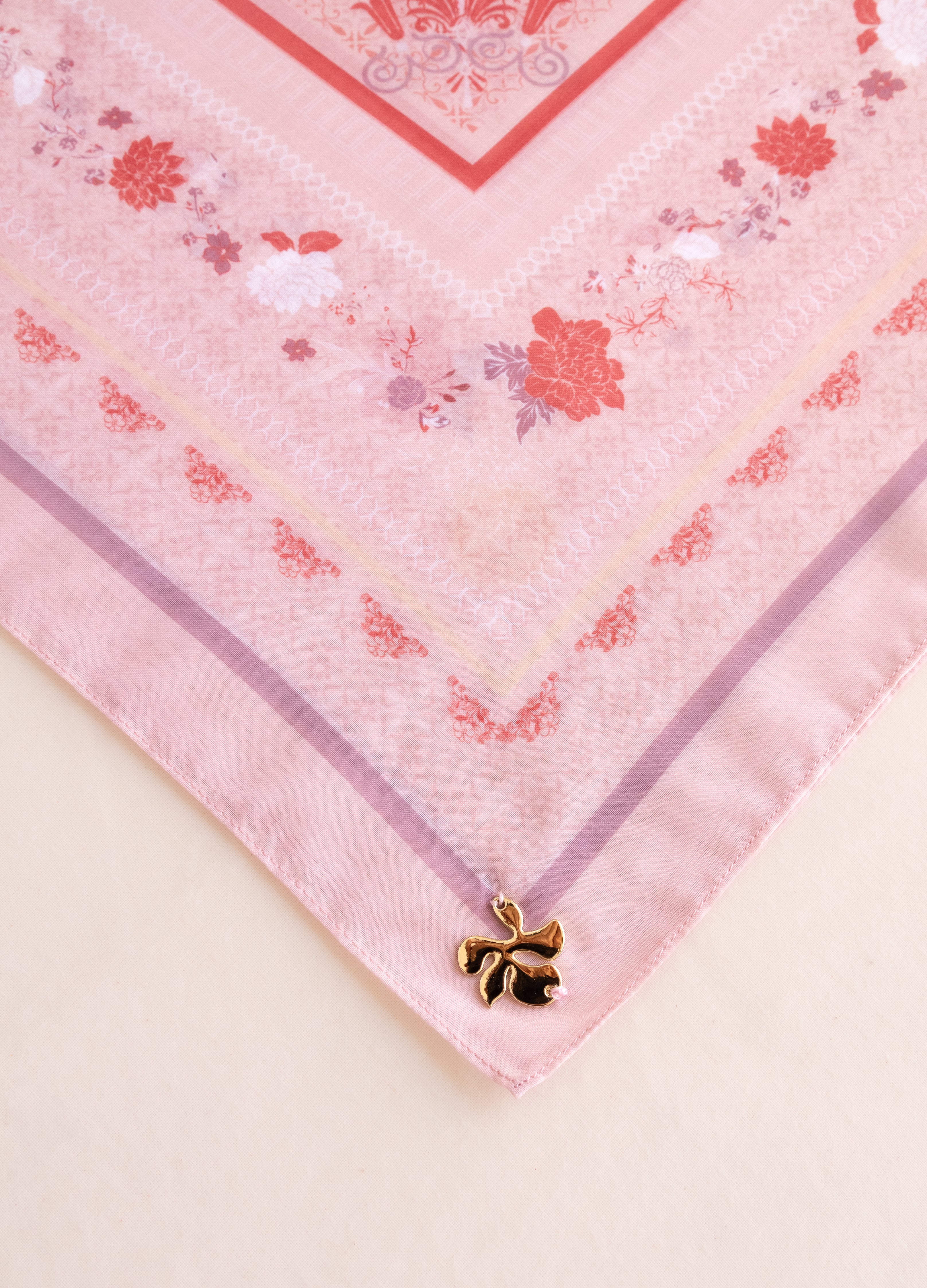 A close-up of a pink patterned scarf or bandana laid flat on a light beige surface, with a small gold bow charm attached near one corner.