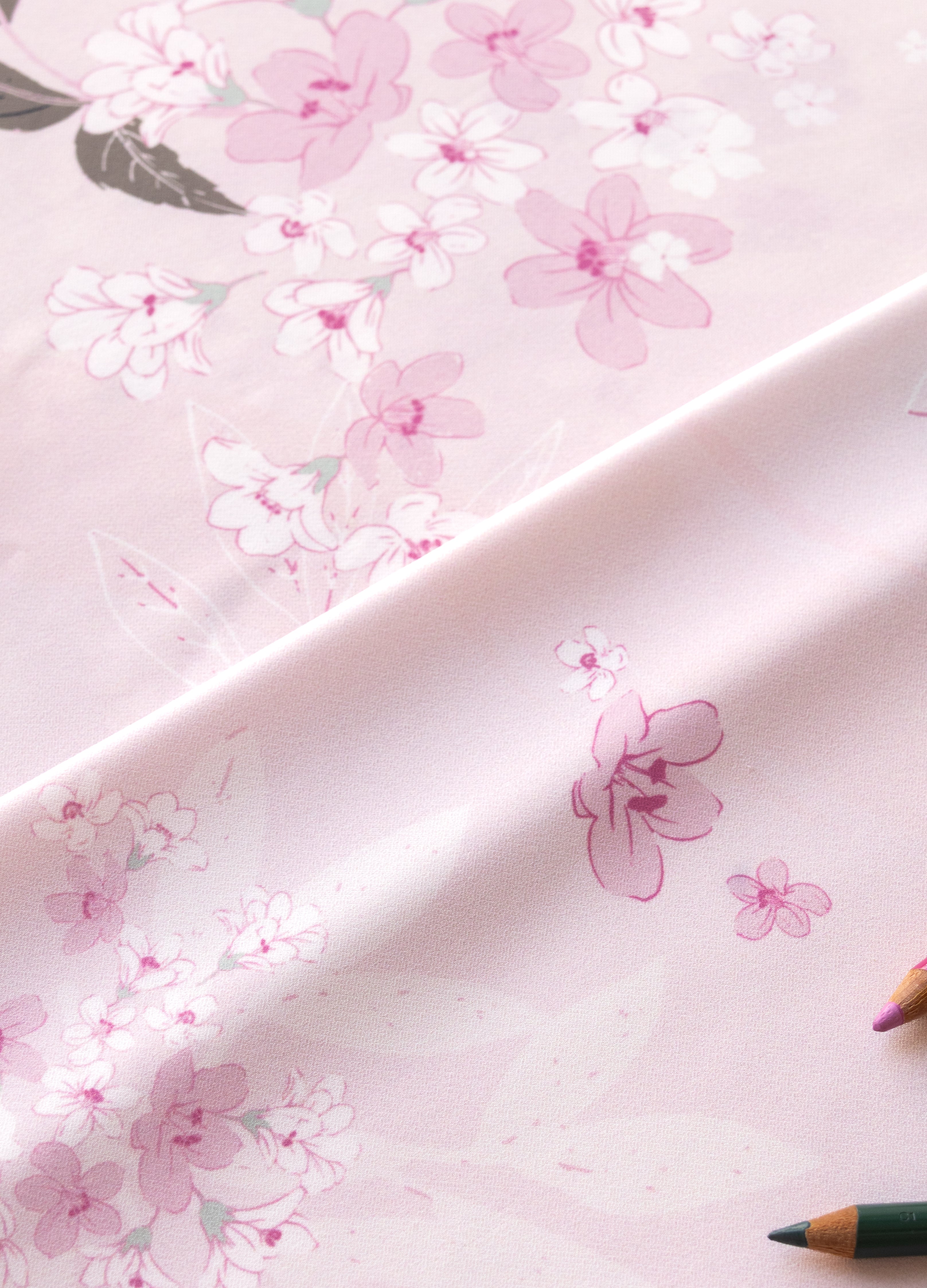 A close-up of a light pink fabric or paper with a delicate floral print in soft pink, white, and muted green, draped diagonally with colored pencils partially visible at the edges.