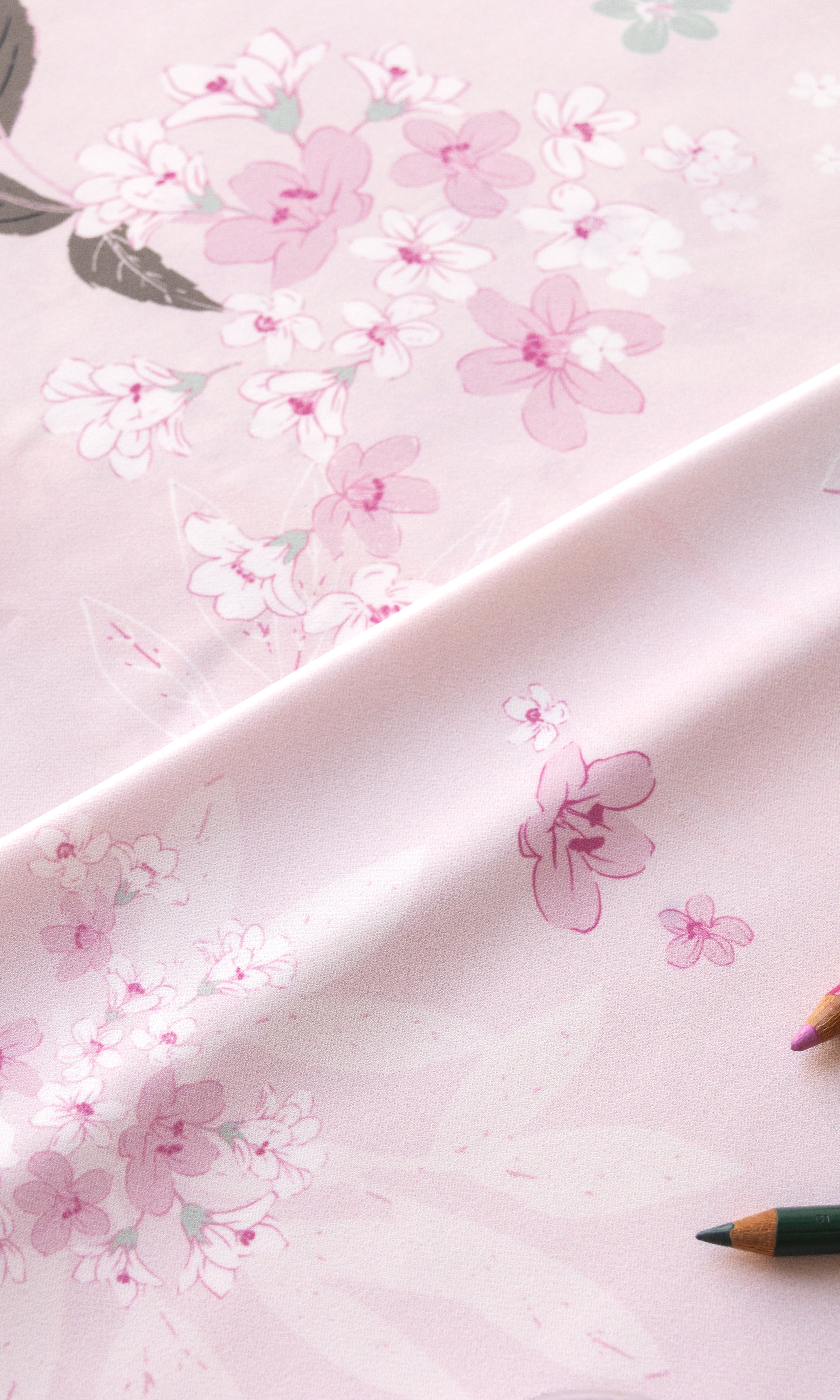 A close-up of a light pink fabric or paper with a delicate floral print in soft pink, white, and muted green, draped diagonally with colored pencils partially visible at the edges.