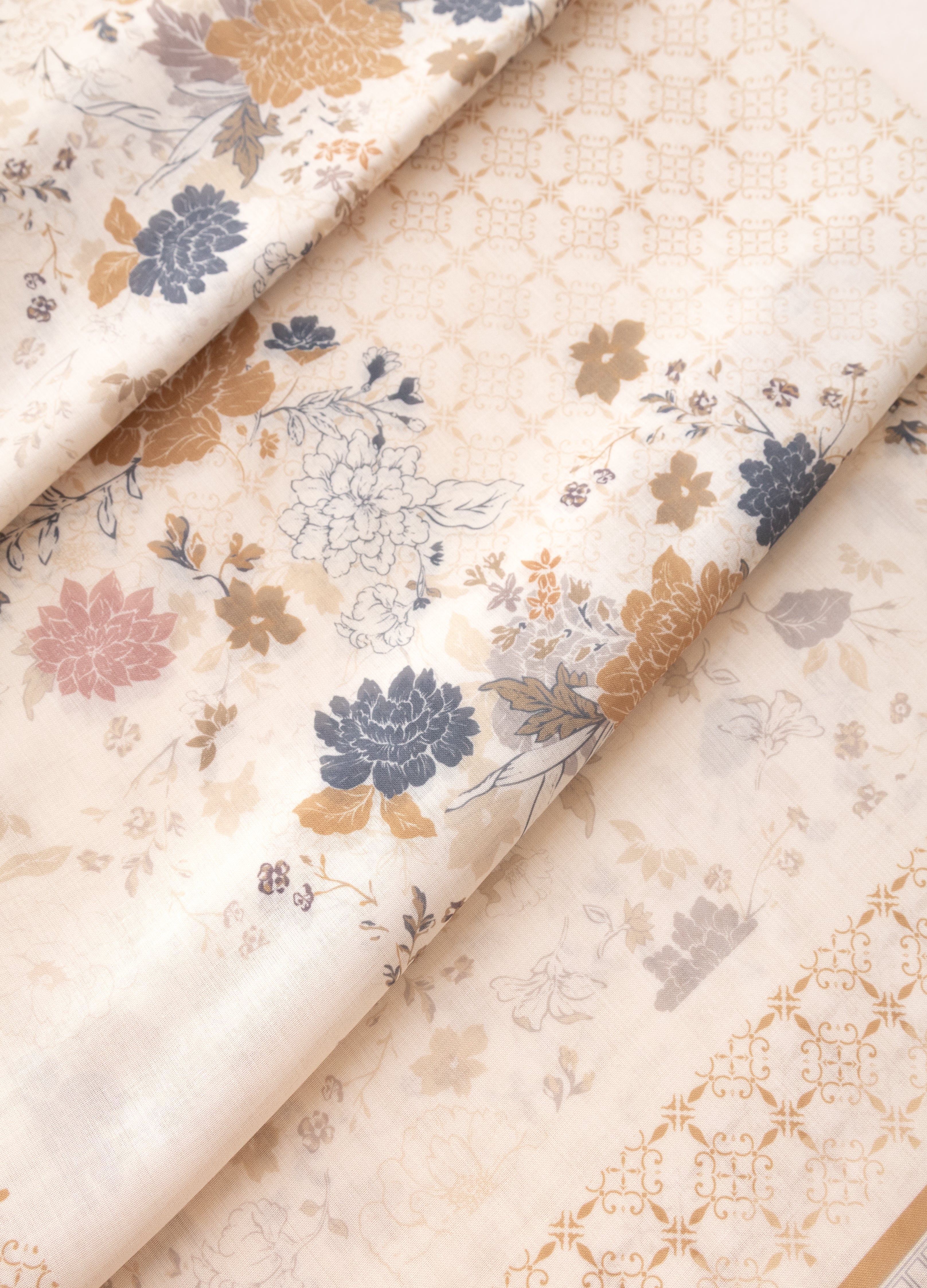 A folded piece of cream-colored fabric with a traditional floral print in muted blue, gold, and pink, featuring a matching gold geometric border.