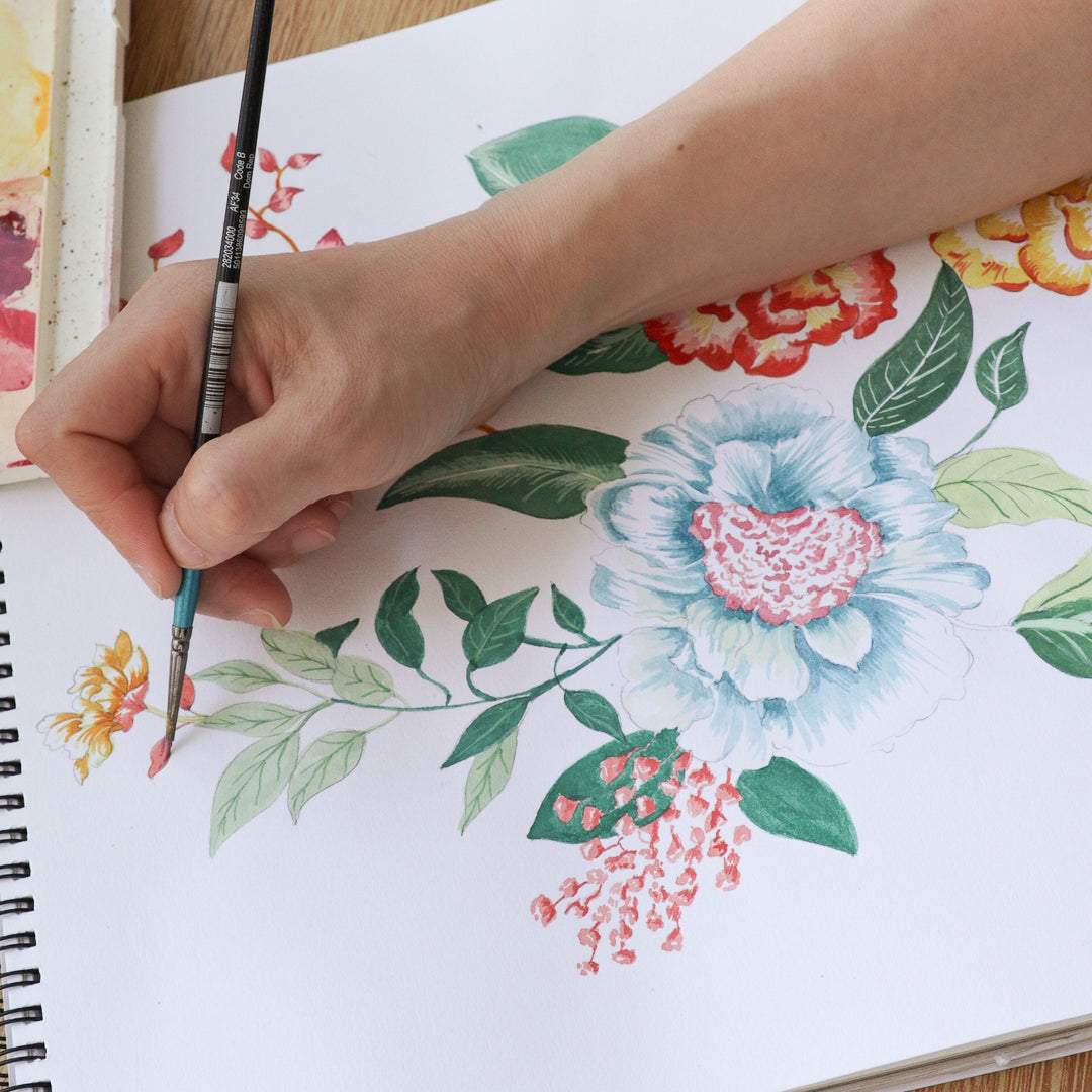 A close-up of a hand holding a paintbrush and painting a floral illustration in a spiral-bound sketchbook. Watercolor paints, brushes, and a palette are arranged on a wooden table.
