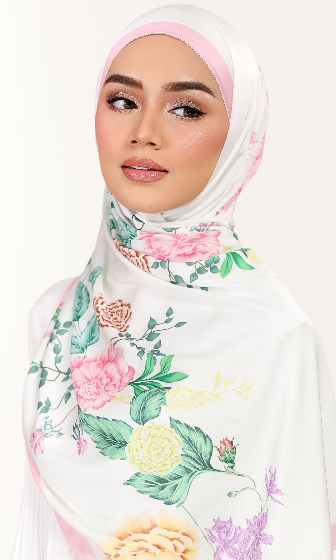 A woman wearing a white hijab with a pastel pink undercap and a colorful floral print featuring pink, yellow, and green roses and leaves, set against a plain white background.