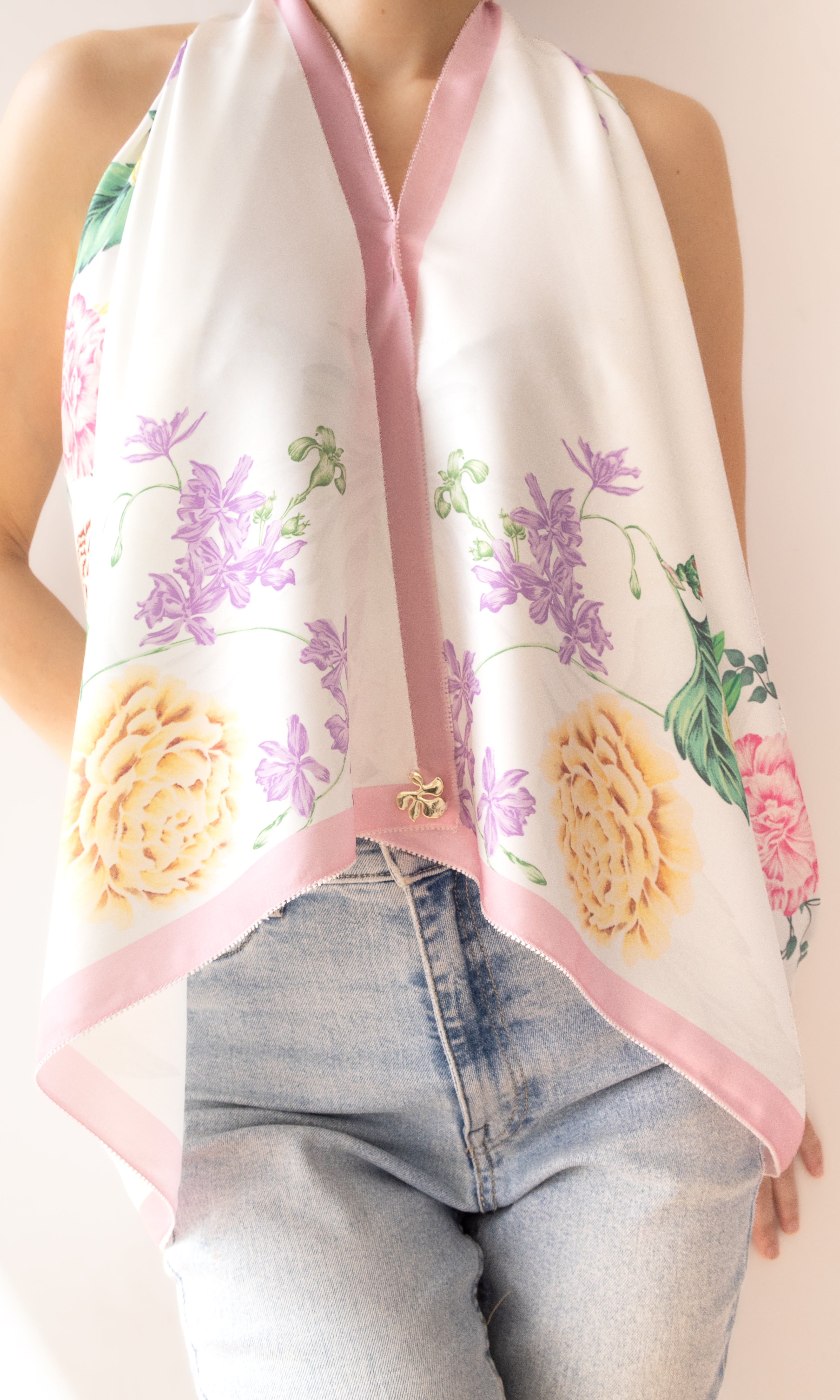 A white halter-style scarf with pink trim and a colorful floral print, worn by a model in light-wash blue jeans against a plain light background.