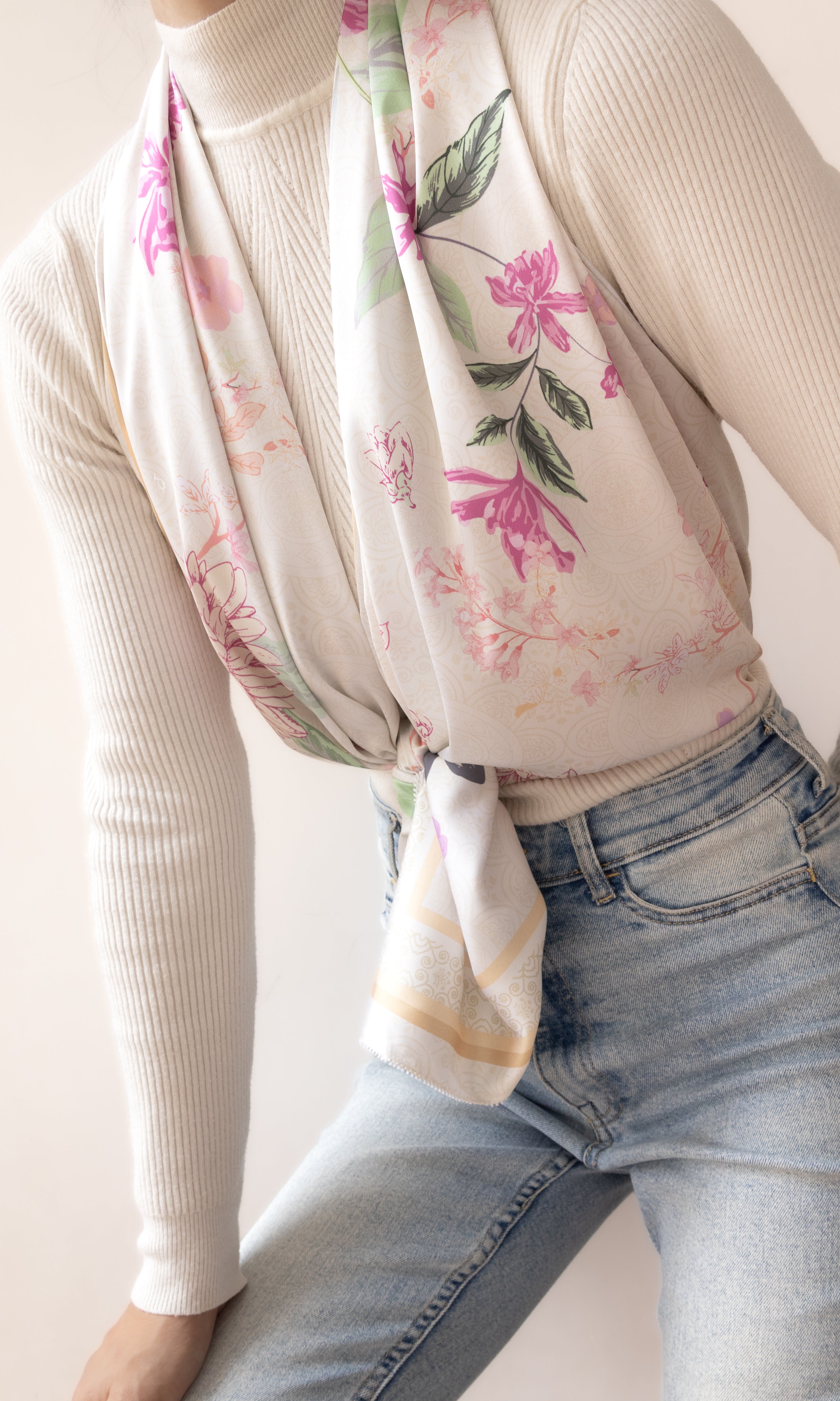 A cream-colored scarf with a pink and green floral print, styled tied around the waist of a model wearing a cream ribbed turtleneck sweater and light blue jeans.