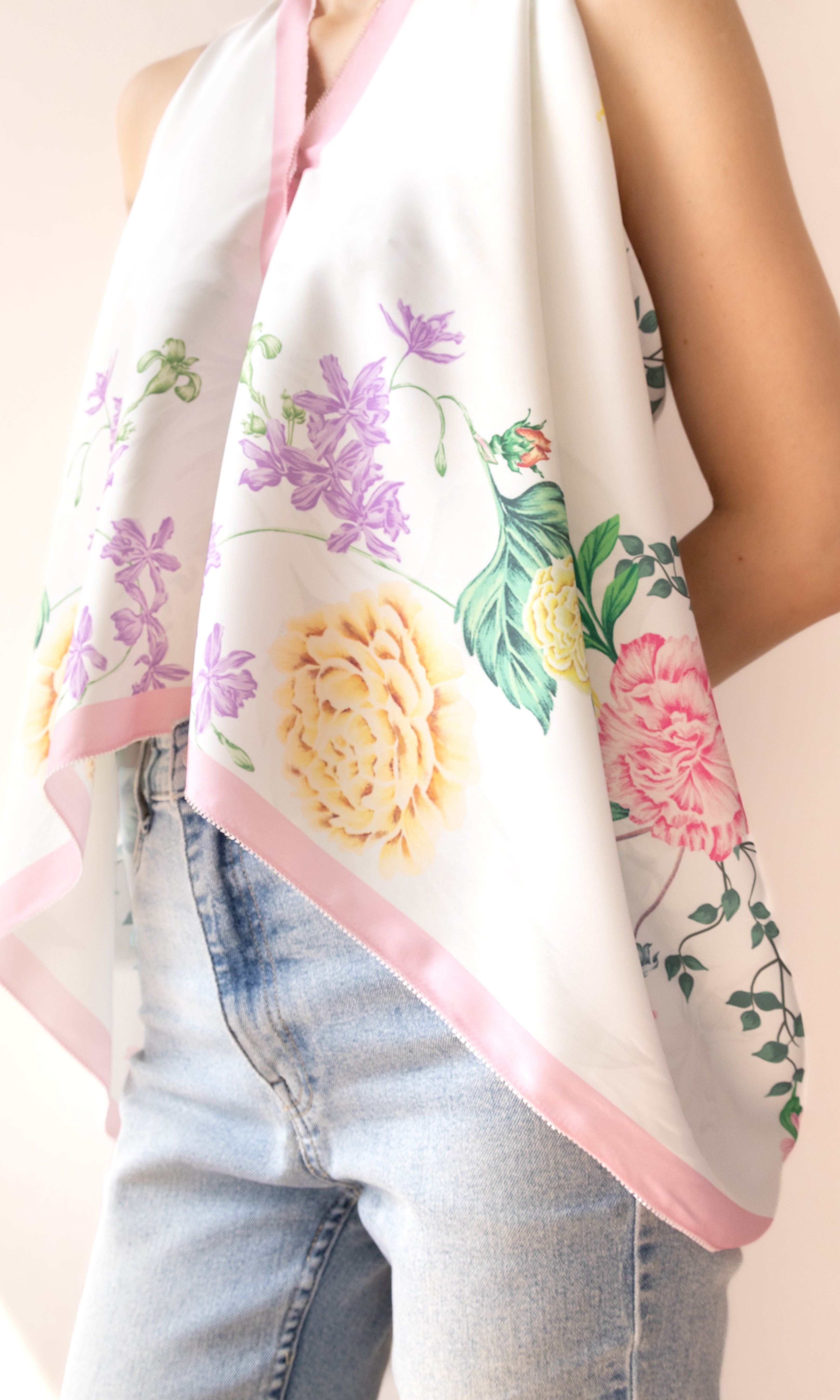 A white halter-style scarf with a pink border and a colorful floral print featuring yellow, pink, and purple flowers with green leaves, worn by a model in light-wash blue jeans.