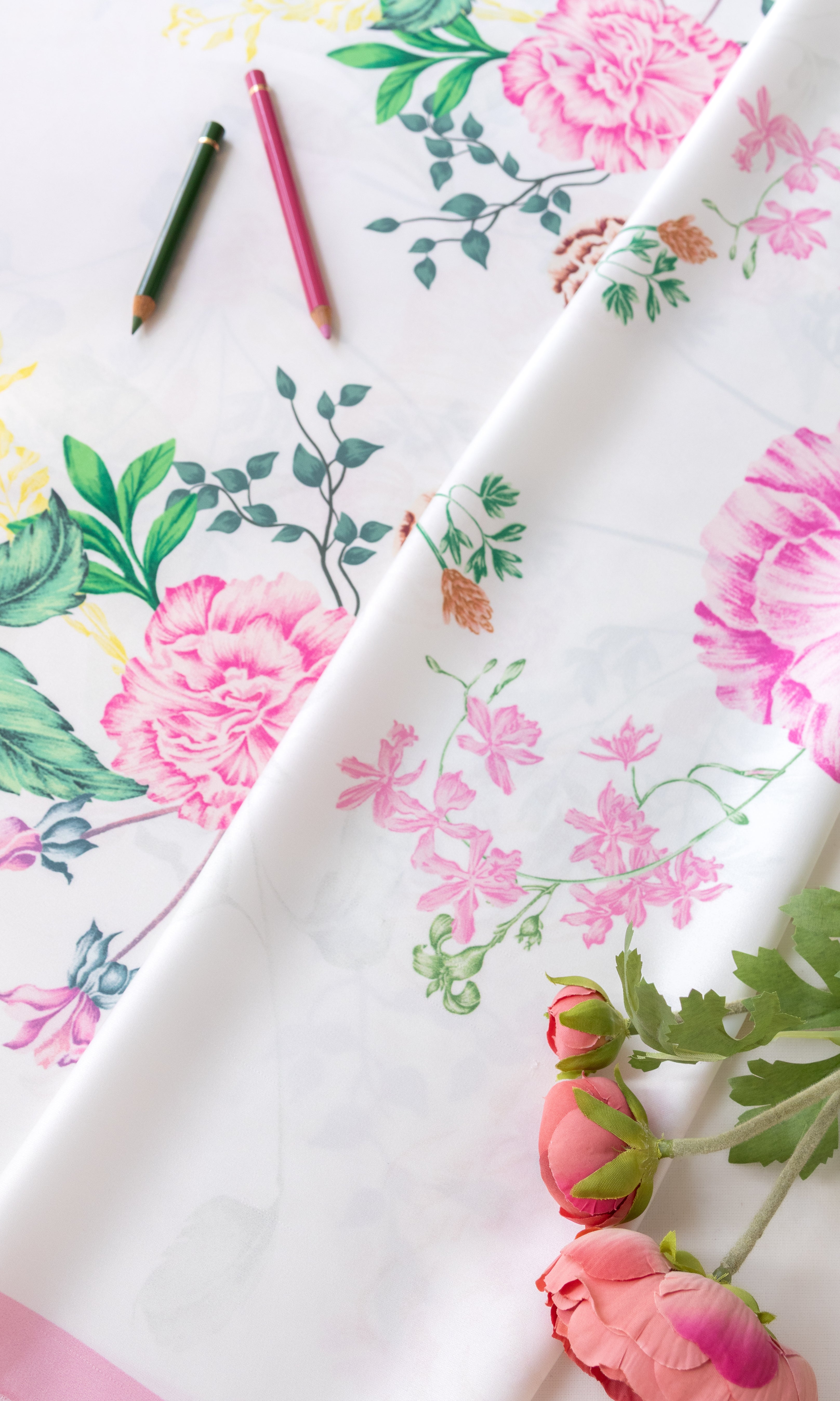 A folded white fabric with a pink and green floral print, styled with two colored pencils and pink flowers on a white background.