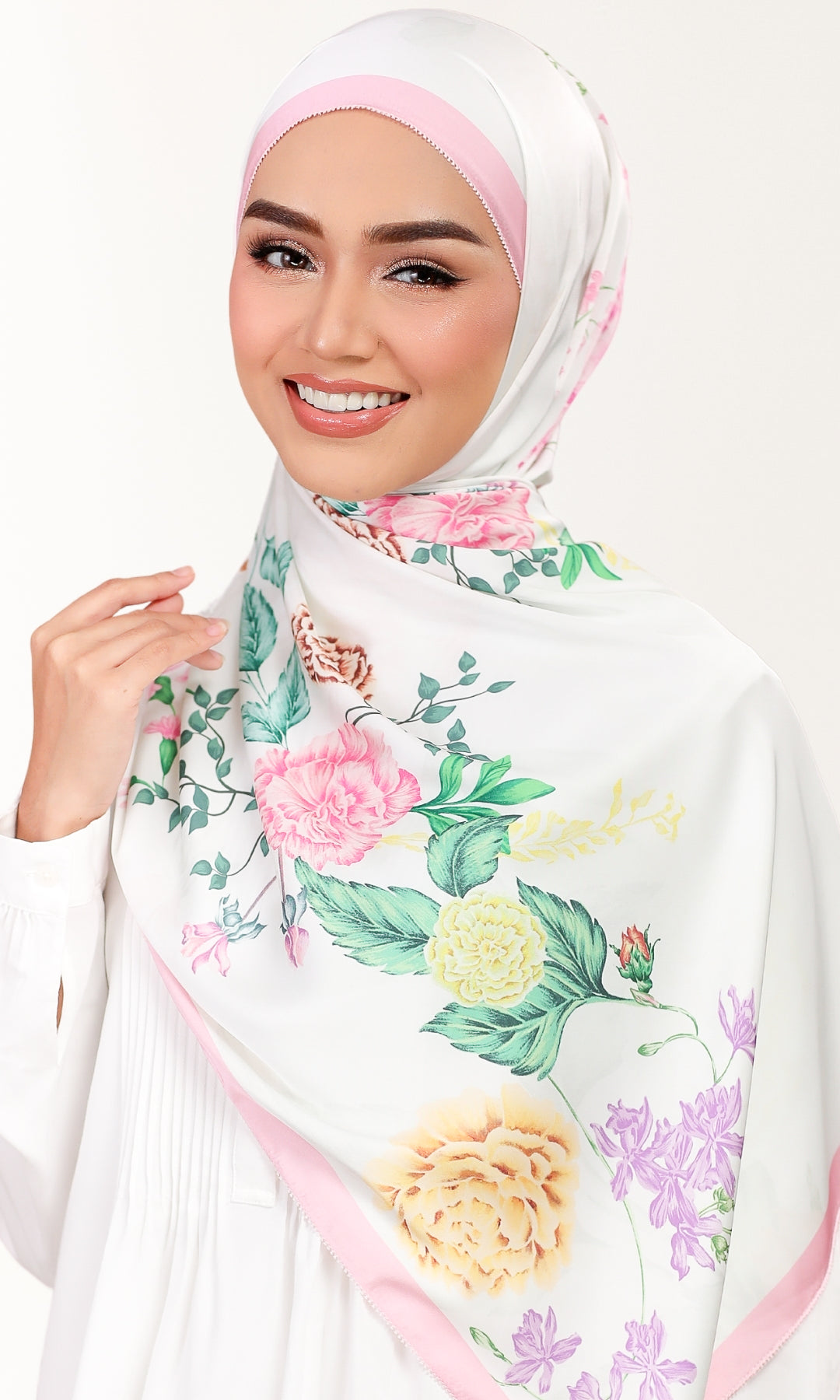 A woman wearing a white hijab with a pink band and a colorful floral print featuring pink, yellow, and purple flowers with green leaves, set against a plain white background.