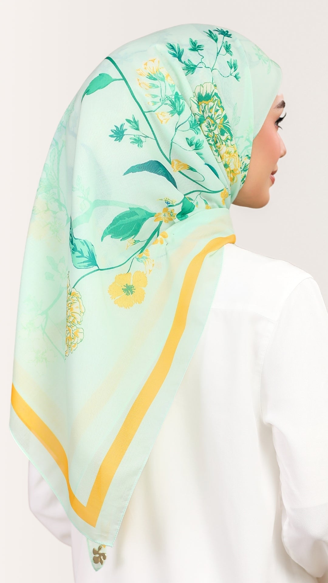 A woman shown from the back wearing a mint green hijab with a yellow and green floral print and a yellow border, paired with a white top against a light beige background.