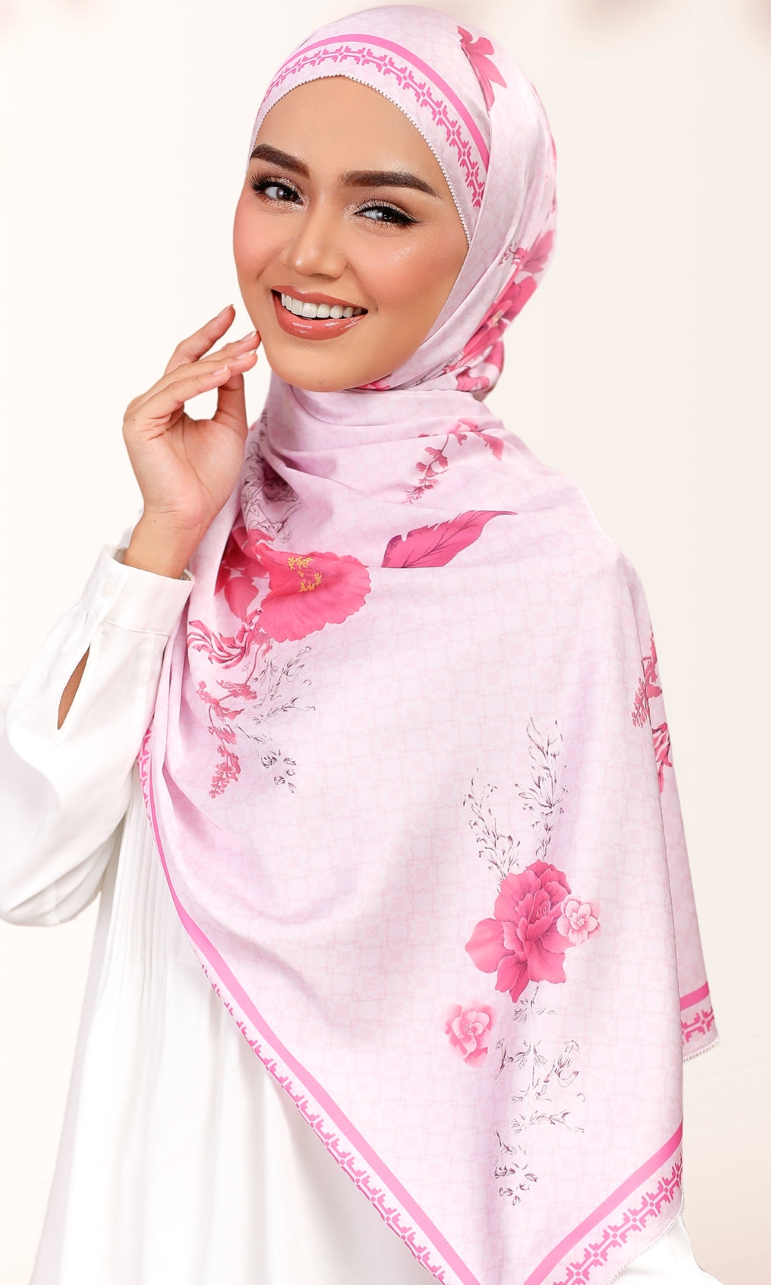 A smiling woman wearing a light pink hijab with a darker pink floral print and delicate border trim, paired with a white long-sleeve top against a soft off-white background.