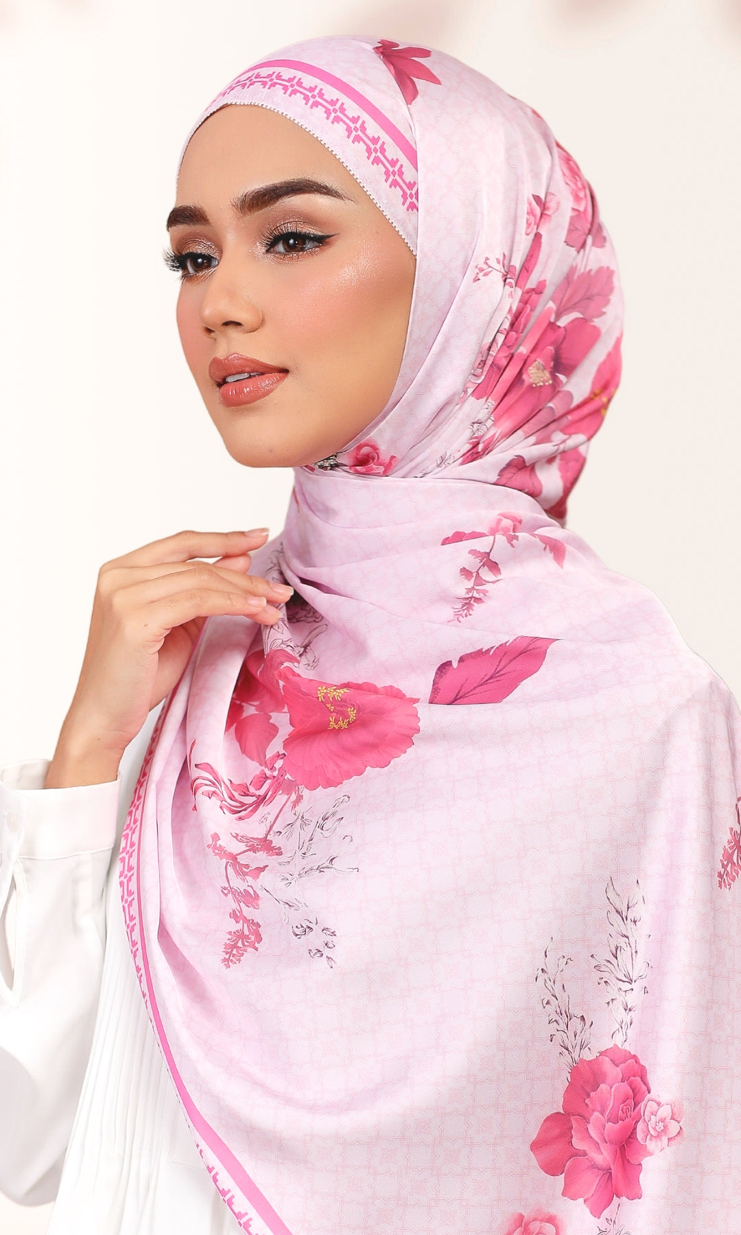 A woman wearing a light pink hijab with a vibrant pink floral print and delicate embroidered trim along the edges, set against a soft, light beige background.