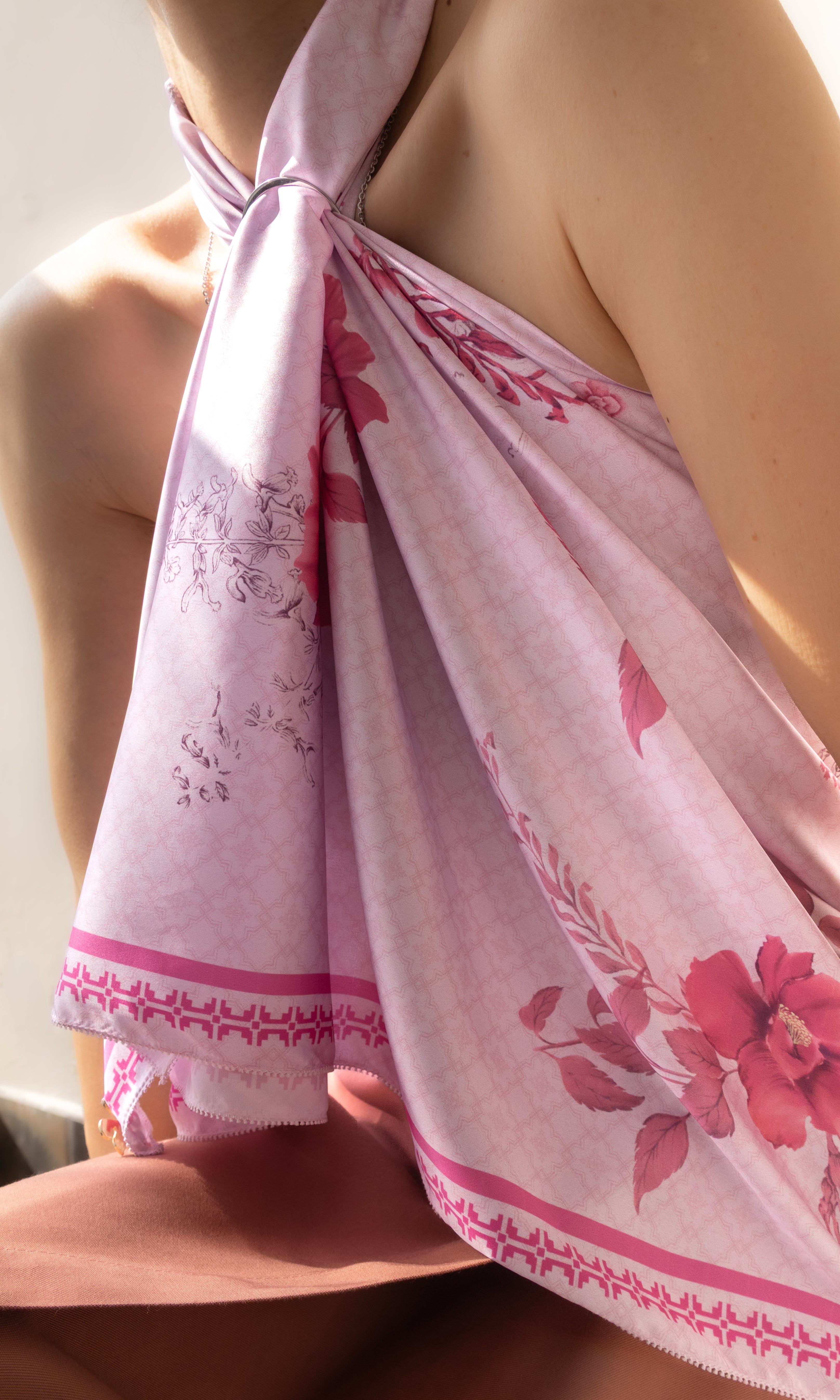 A close-up of a person wearing a light pink halter-style wrap or scarf with a floral print and decorative border, tied around the neck and draped over the shoulder.