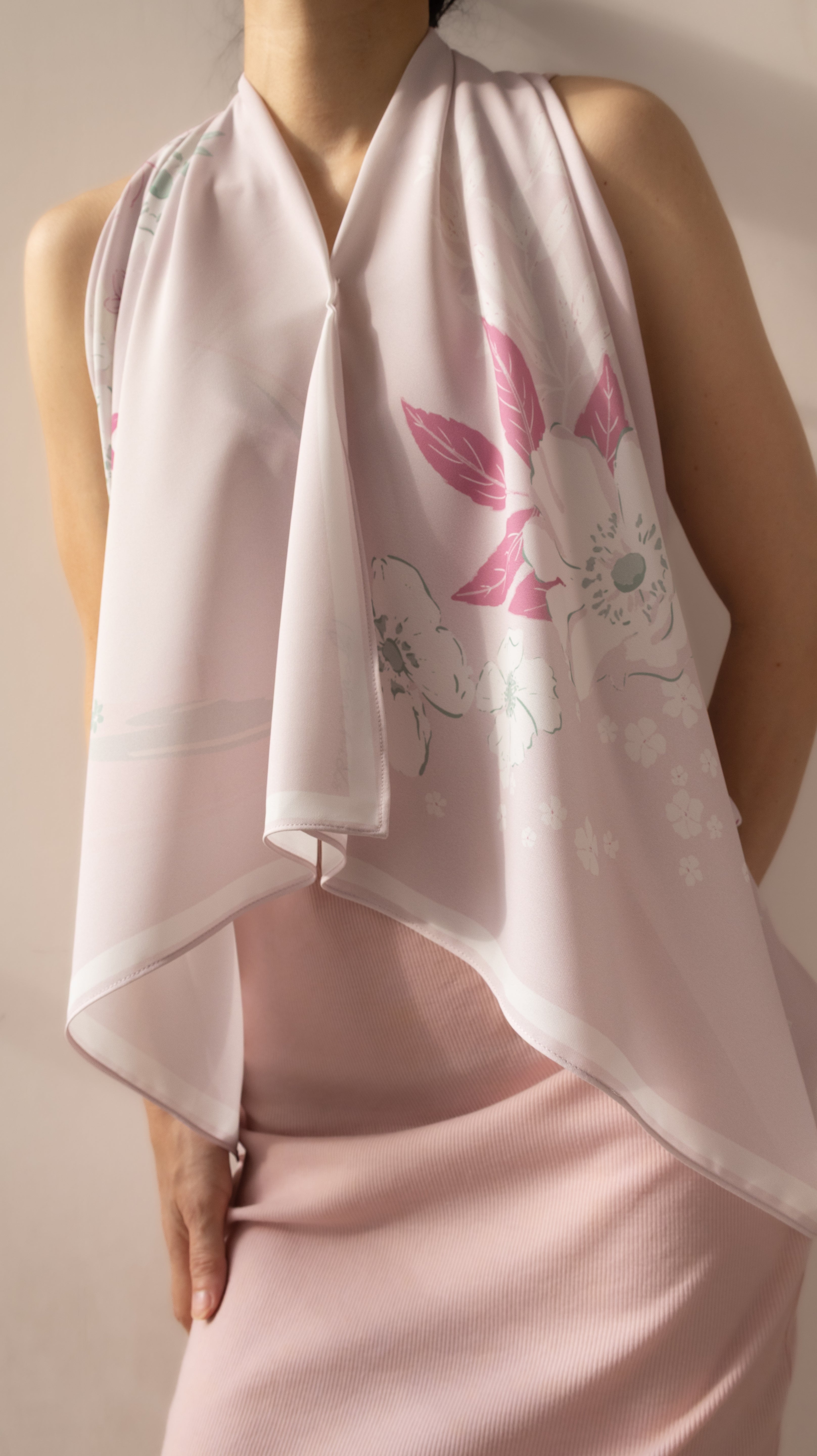 A close-up of a model wearing a light pink halter-style scarf with a floral print in pink and white, paired with a matching light pink satin dress.