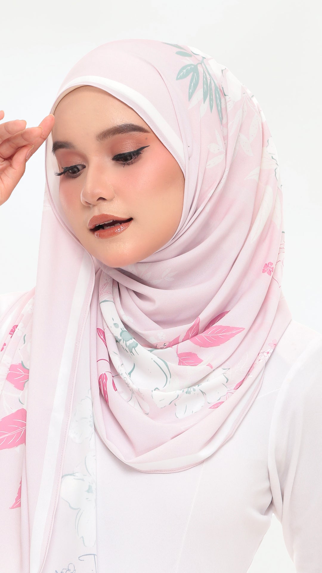 A woman wearing a light pink hijab with a delicate green and pink floral print, styled against a clean white background.