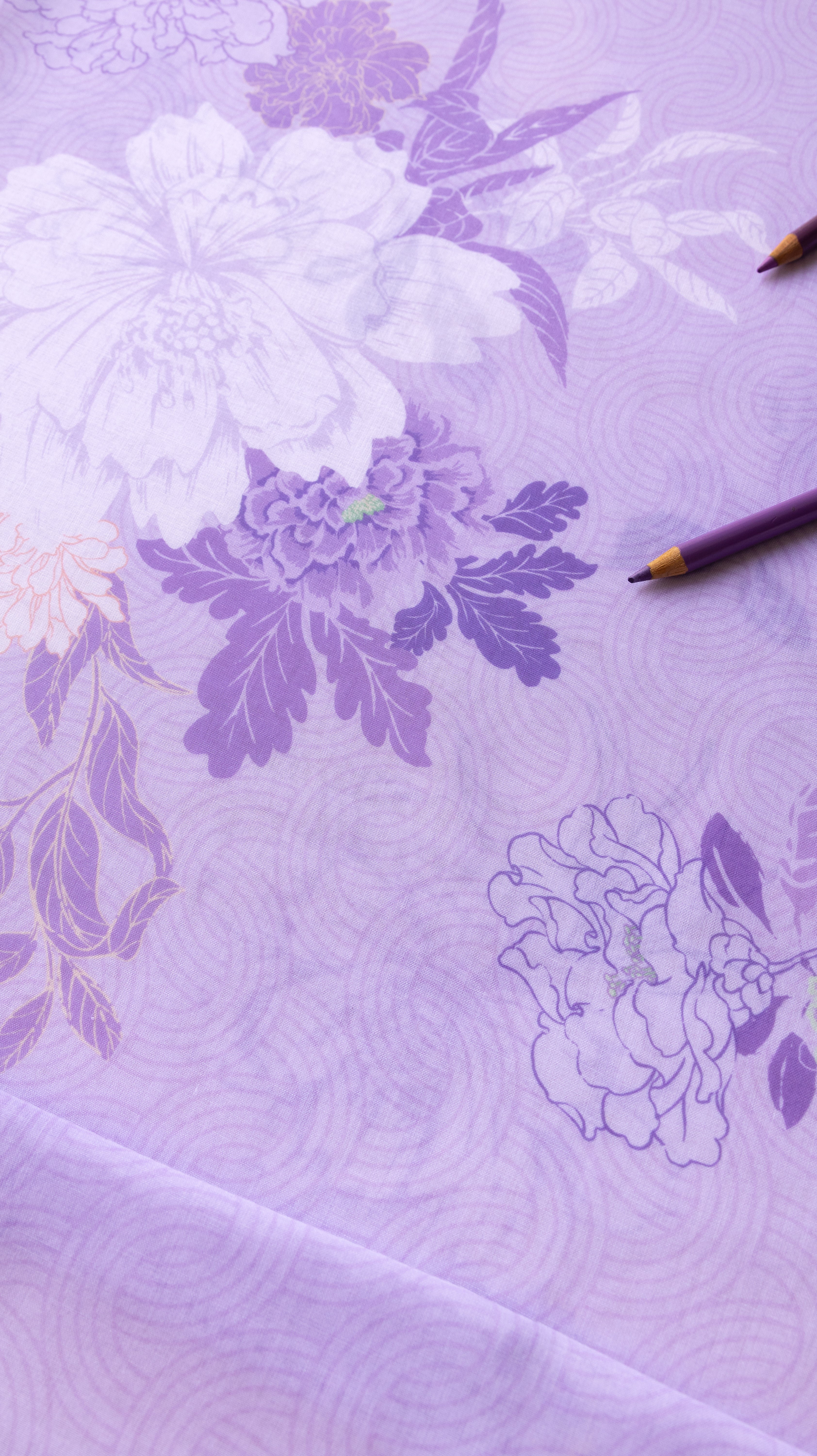 A close-up of a lavender purple fabric with a floral pattern in shades of white and darker purple, featuring a subtle wave texture. Two colored pencils are placed on the surface.
