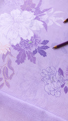 A close-up of a lavender purple fabric with a floral pattern in shades of white and darker purple, featuring a subtle wave texture. Two colored pencils are placed on the surface.