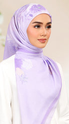 A woman wearing a light lavender purple hijab with a subtle floral pattern, styled with a white top against a soft beige background.