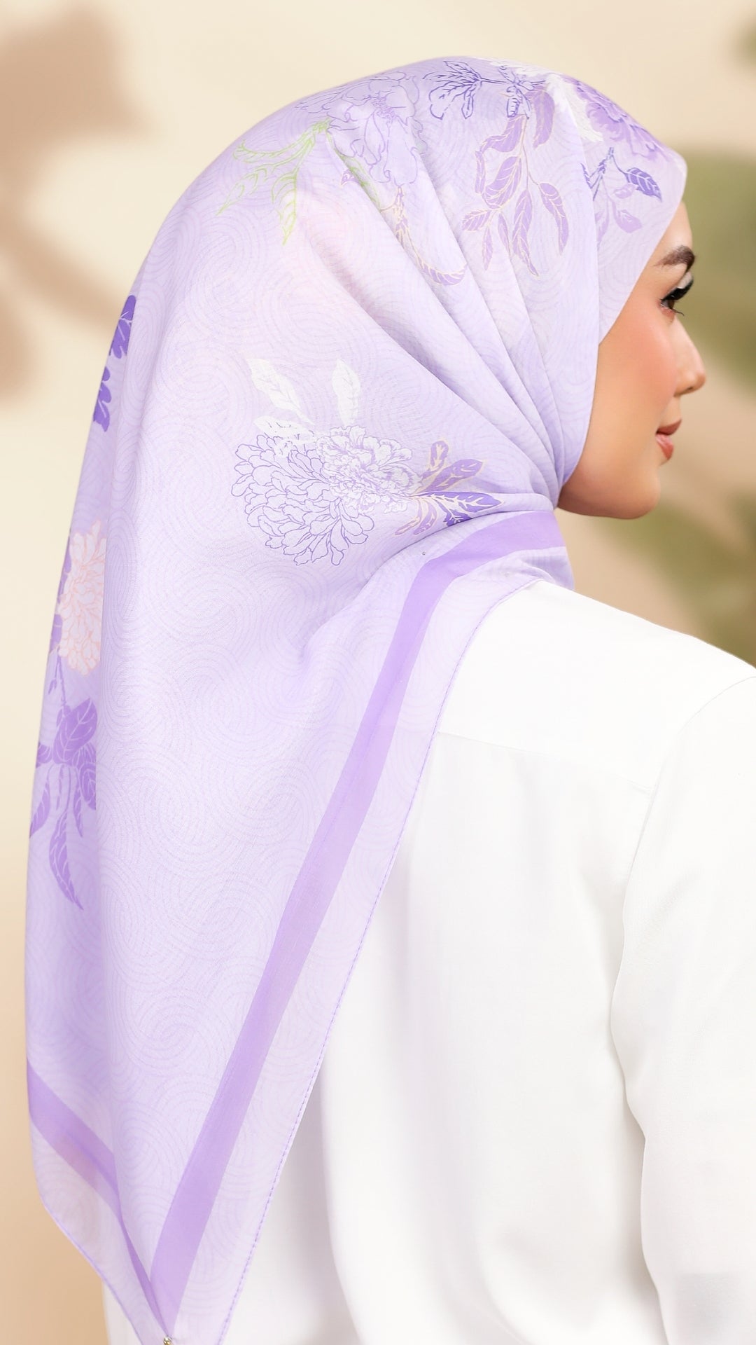 A woman wearing a light lavender purple hijab with a subtle floral pattern and solid border, paired with a white top. The photo is taken from a side-back angle against a soft, warm beige background.