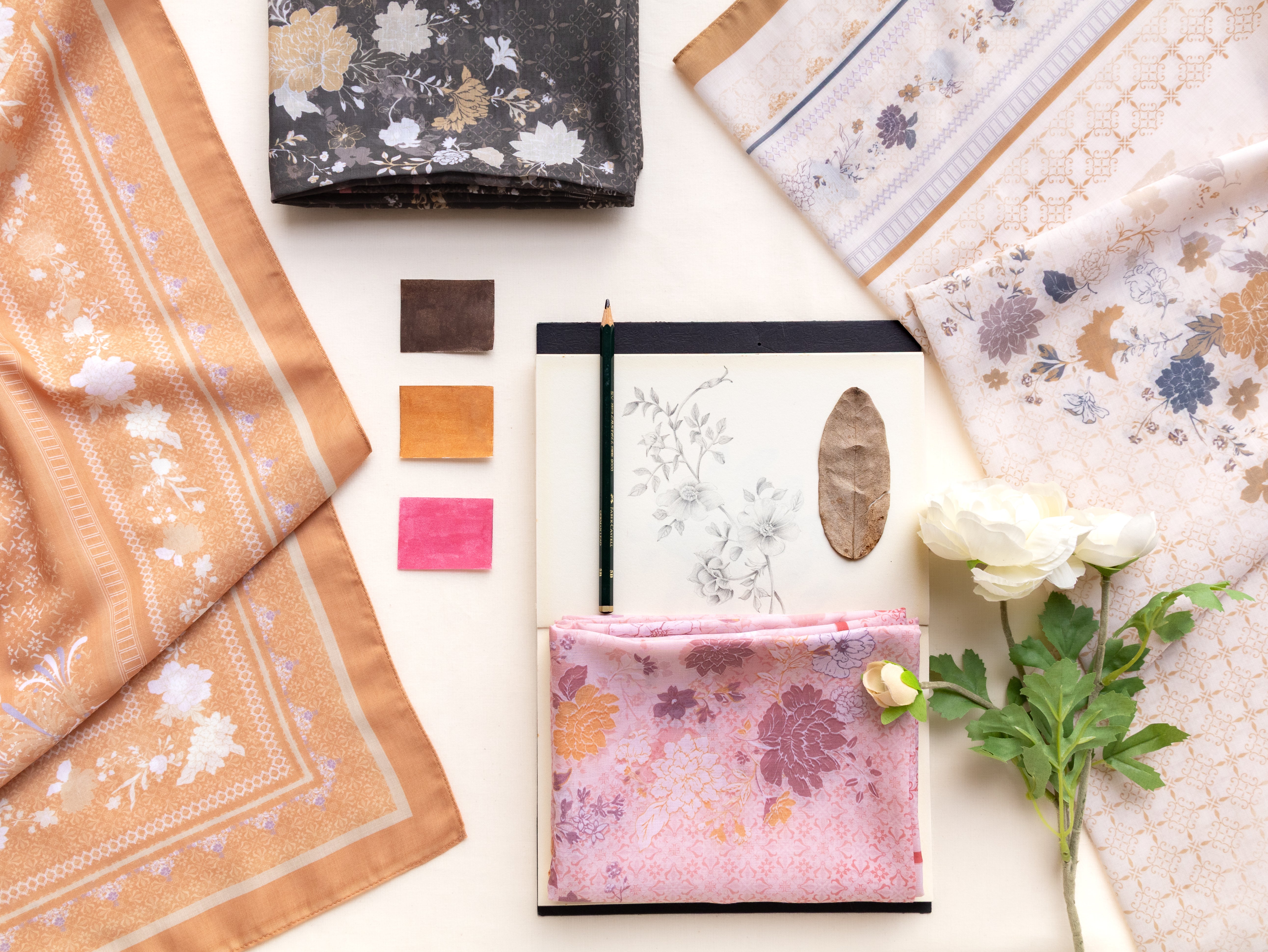 A styled flat-lay of multiple silk scarves with floral and geometric patterns in soft peach, pink, beige, and gray tones, arranged around a notebook and fabric swatches on a cream background.
