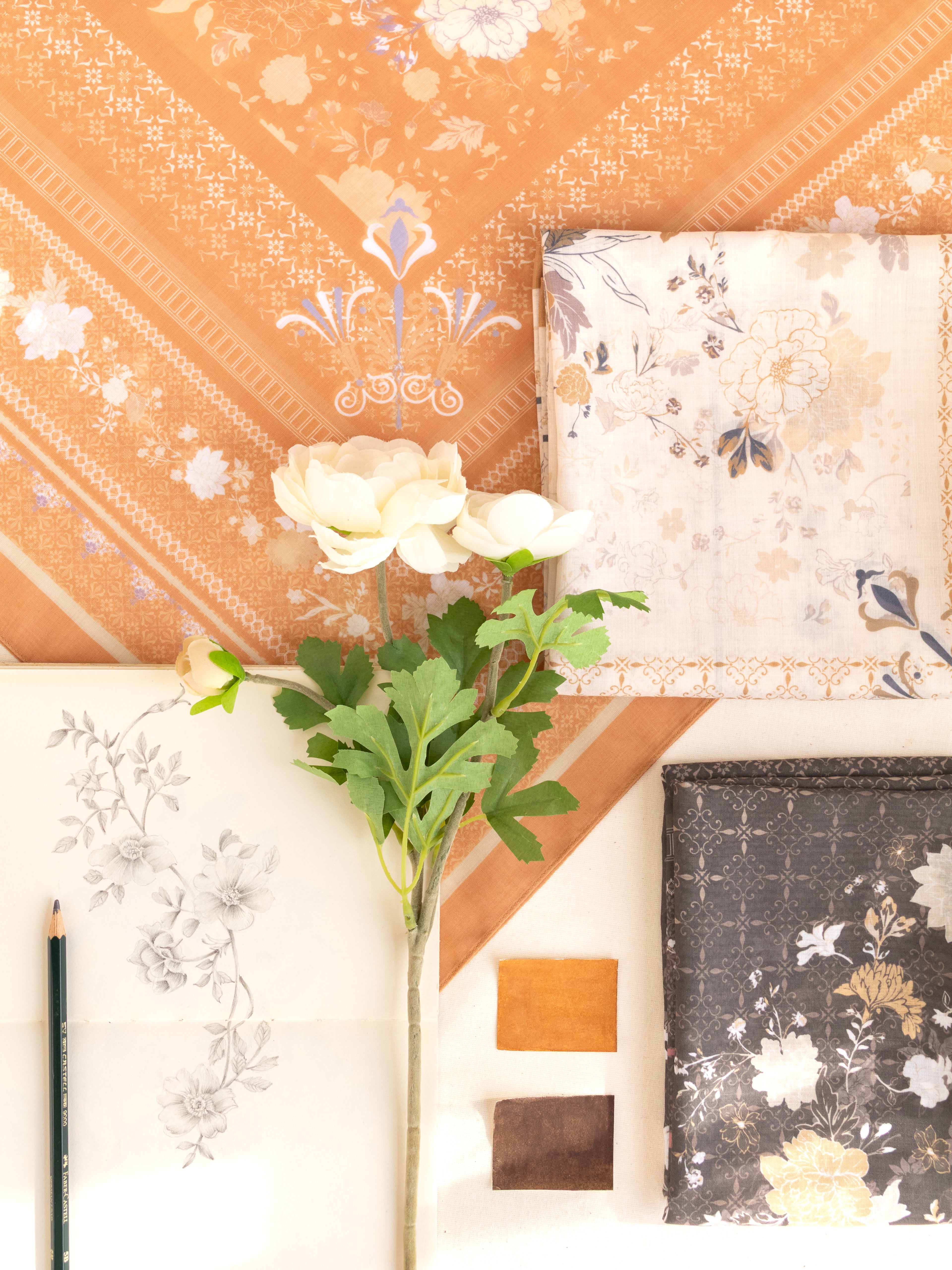 A styled flat-lay showcasing various fabric or wallpaper swatches with floral and geometric patterns in warm tones of orange, beige, white, and dark gray, arranged around a single white rose stem on a light background.