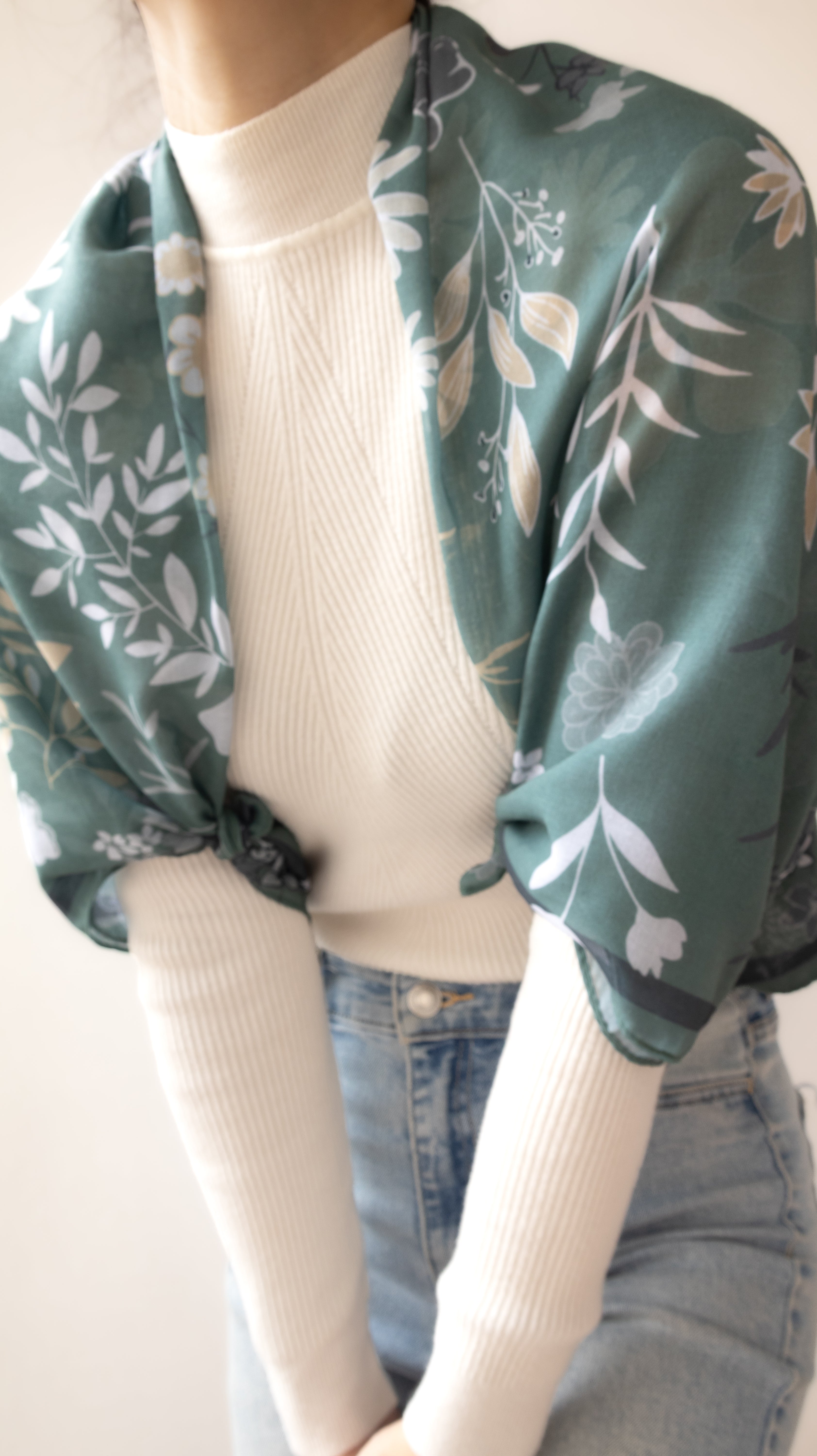 A close-up of a person wearing a sage green floral silk scarf draped over a cream ribbed turtleneck sweater and light blue jeans.