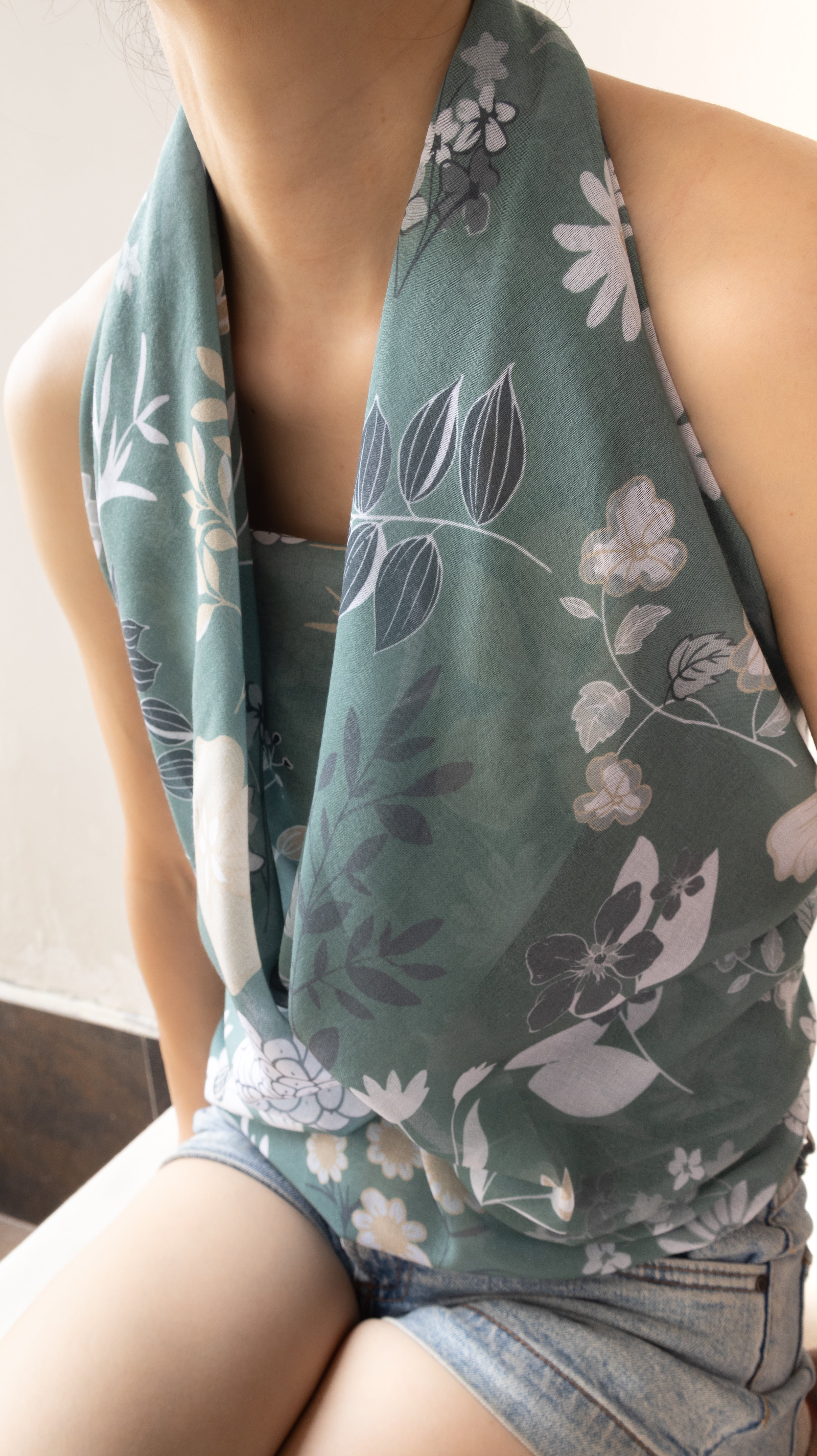 A close-up of a person wearing a sage green halter-style top or scarf with a white and gray floral print, paired with denim shorts.