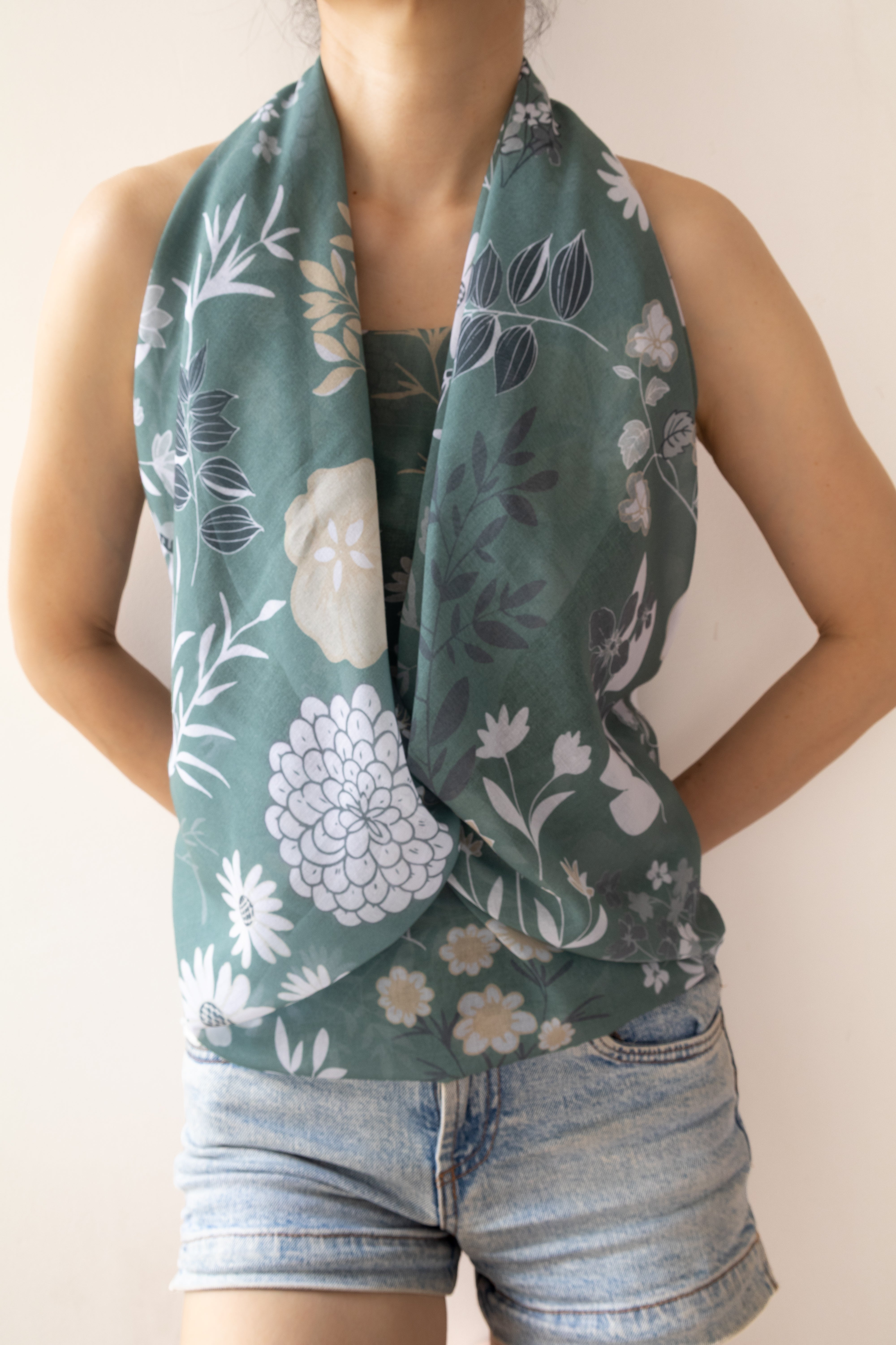 A woman wearing a sage green floral scarf draped around her neck, paired with light-wash denim shorts, against a plain beige background.