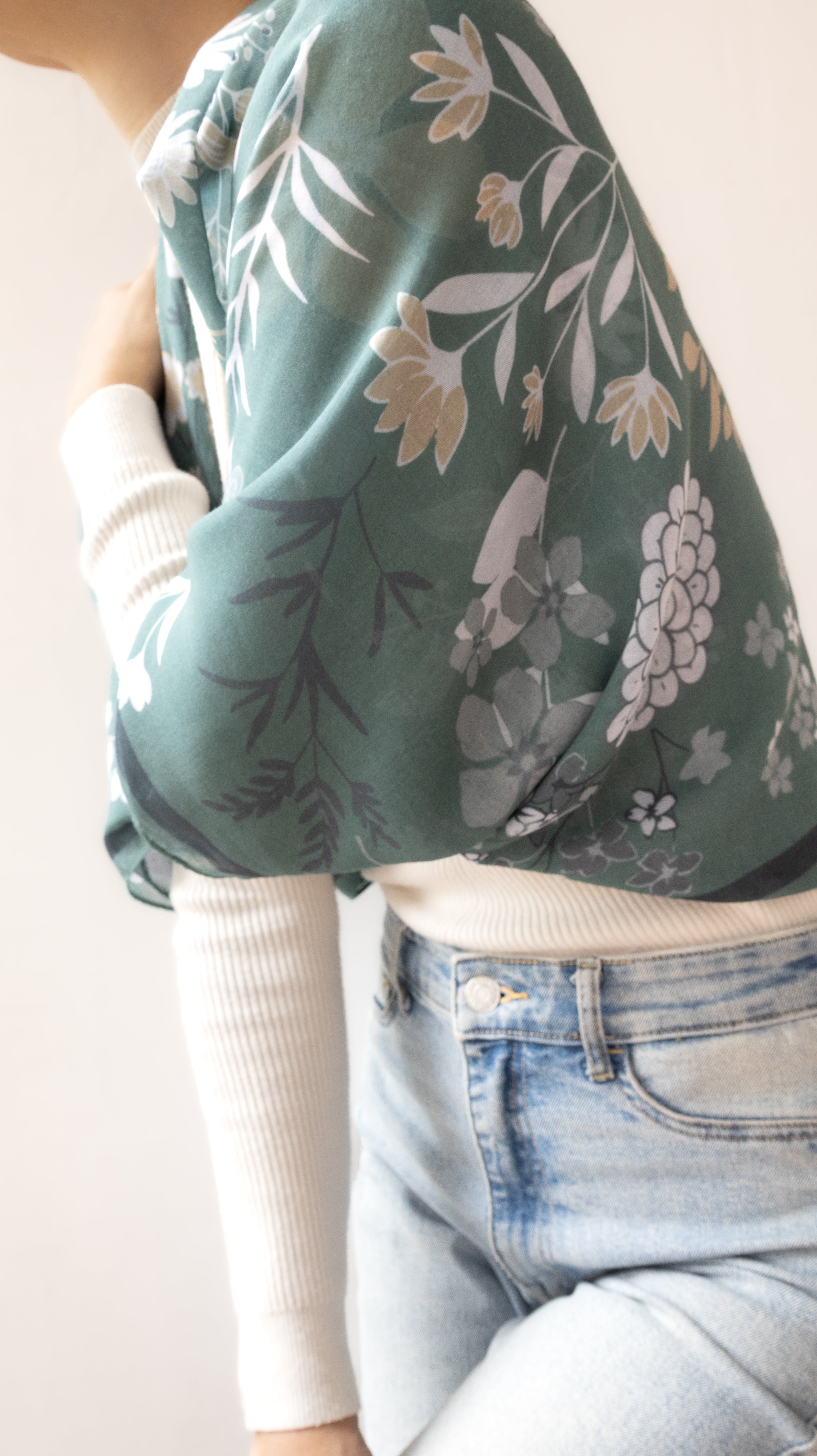 A close-up of a person wearing a sage green floral long-sleeve top over a white ribbed long-sleeve shirt, paired with light blue jeans.