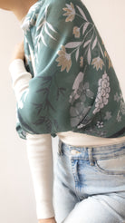 A close-up of a person wearing a sage green floral long-sleeve top over a white ribbed long-sleeve shirt, paired with light blue jeans.