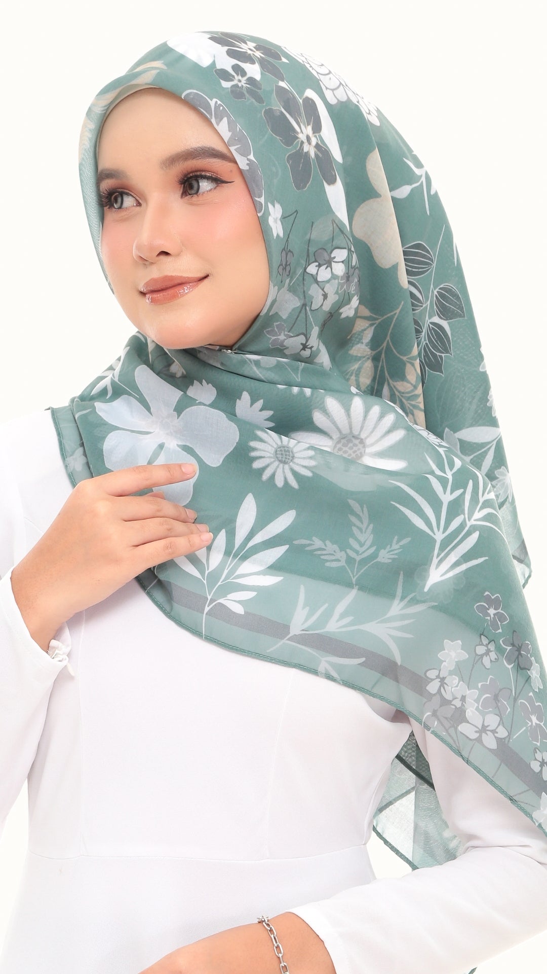 A woman wearing a sage green hijab with a white and gray floral pattern, paired with a white long-sleeve top against a plain white background.