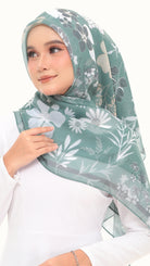 A woman wearing a sage green hijab with a white and gray floral pattern, paired with a white long-sleeve top against a plain white background.