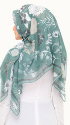 A woman wearing a semi-sheer sage green hijab with a white, gray, and beige floral pattern, styled over a white top against a plain white background.