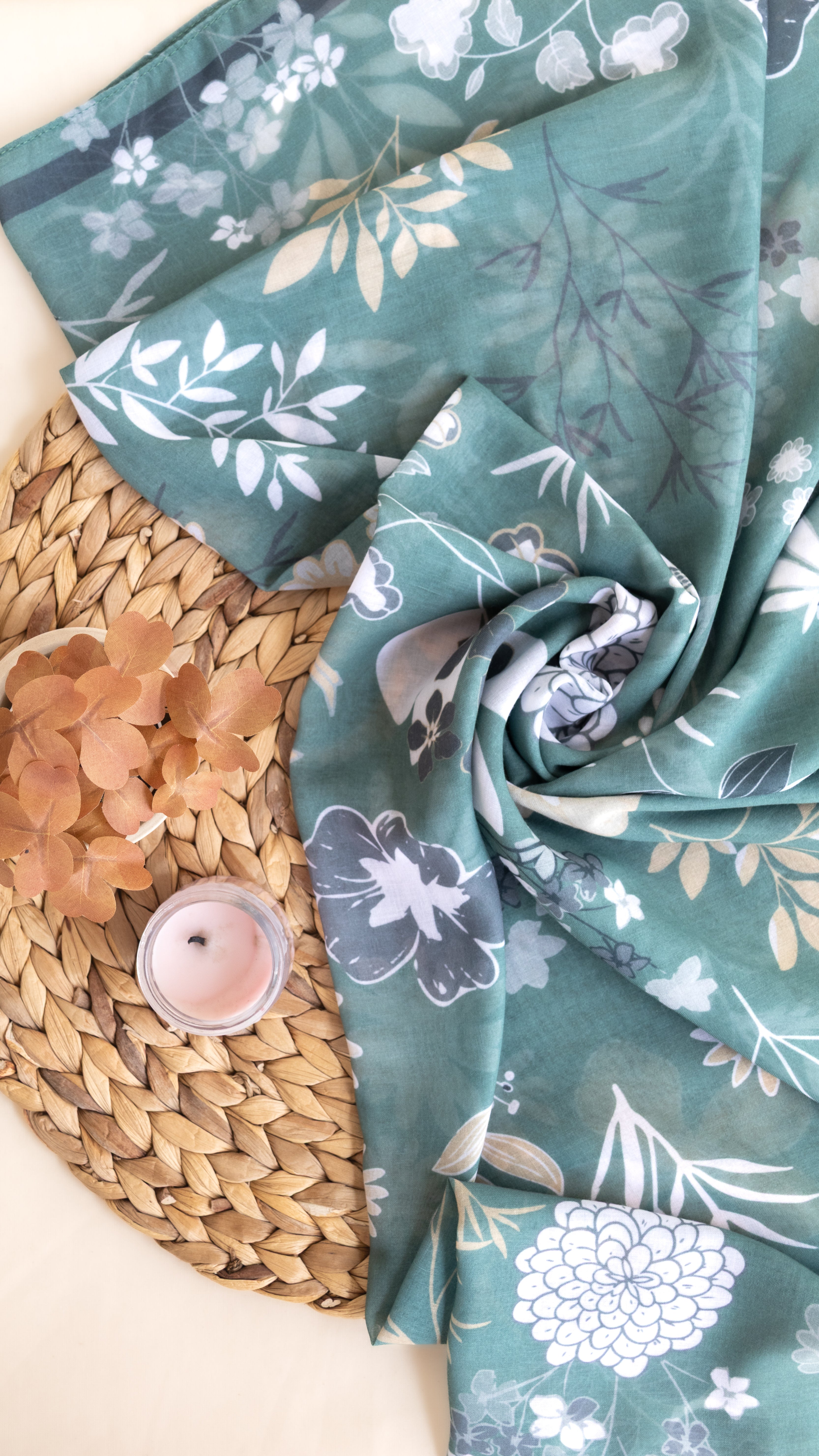 A teal floral scarf with white and gray botanical patterns, draped elegantly over a woven straw mat next to a small lit candle and dried peach petals.