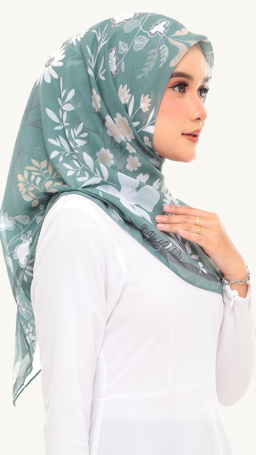 A woman wearing a sage green floral hijab and a white long-sleeve top, shown in a side profile against a plain light background.