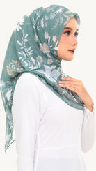 A woman wearing a sage green floral hijab and a white long-sleeve top, shown in a side profile against a plain light background.