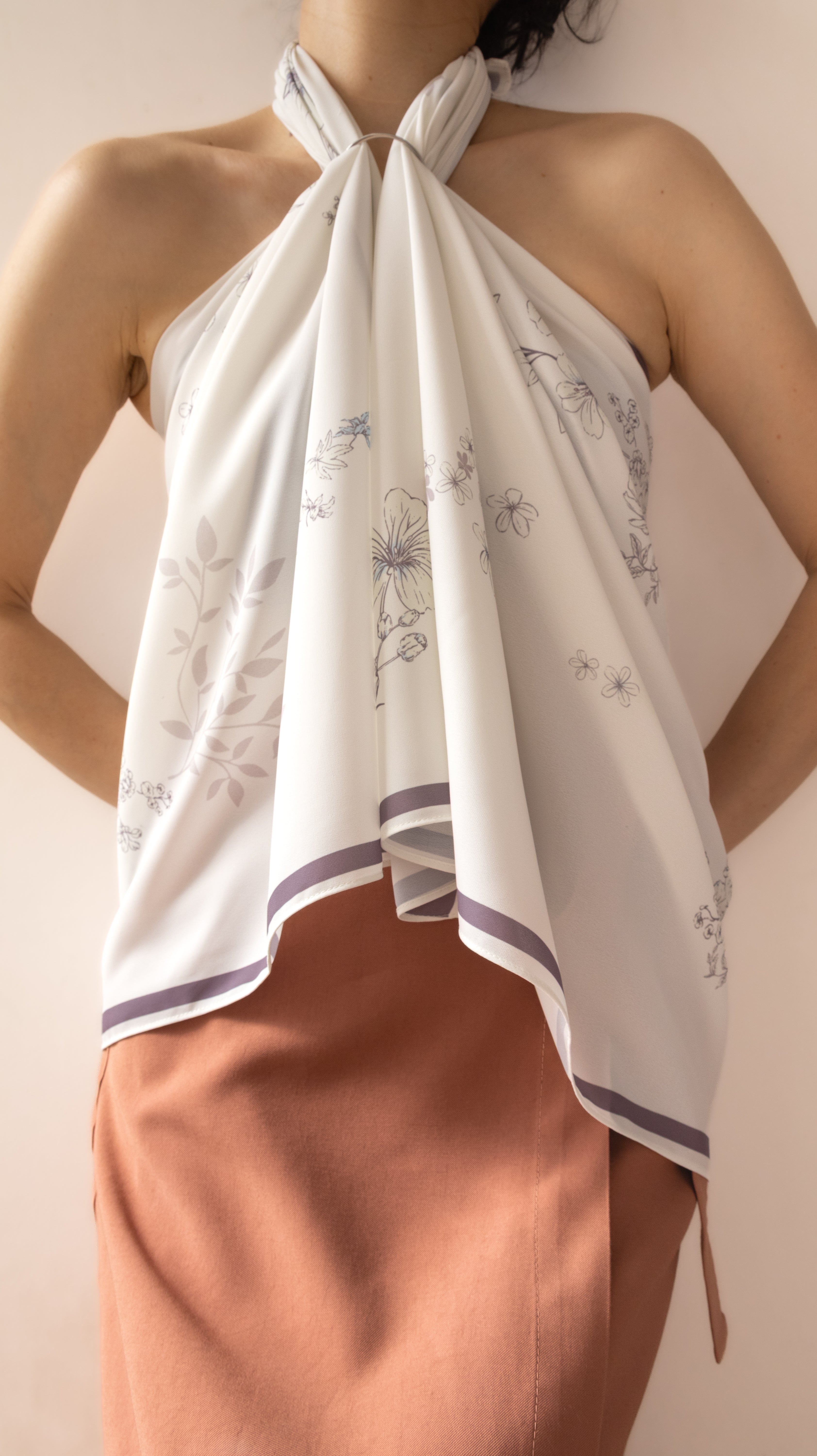 A woman wearing a white halter-neck scarf top with a delicate gray floral pattern and purple trim, paired with a peach-colored skirt, against a soft beige background.
