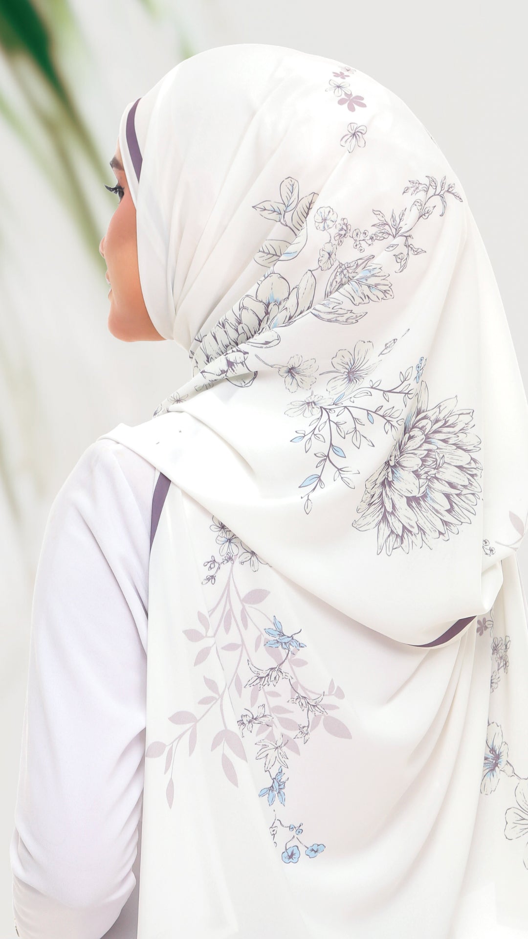 A woman shown from the side wearing a white hijab with a delicate floral print in shades of blue and gray, paired with a white long-sleeve top.