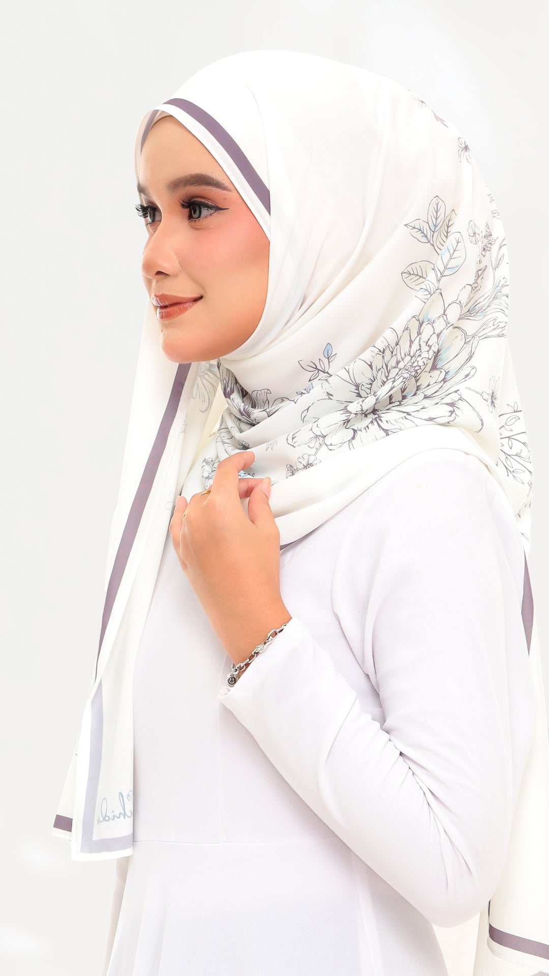 A woman wearing a white hijab with delicate gray floral patterns and thin purple side stripes, paired with a white long-sleeve top against a plain white background.