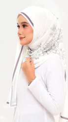 A woman wearing a white hijab with delicate gray floral patterns and thin purple side stripes, paired with a white long-sleeve top against a plain white background.