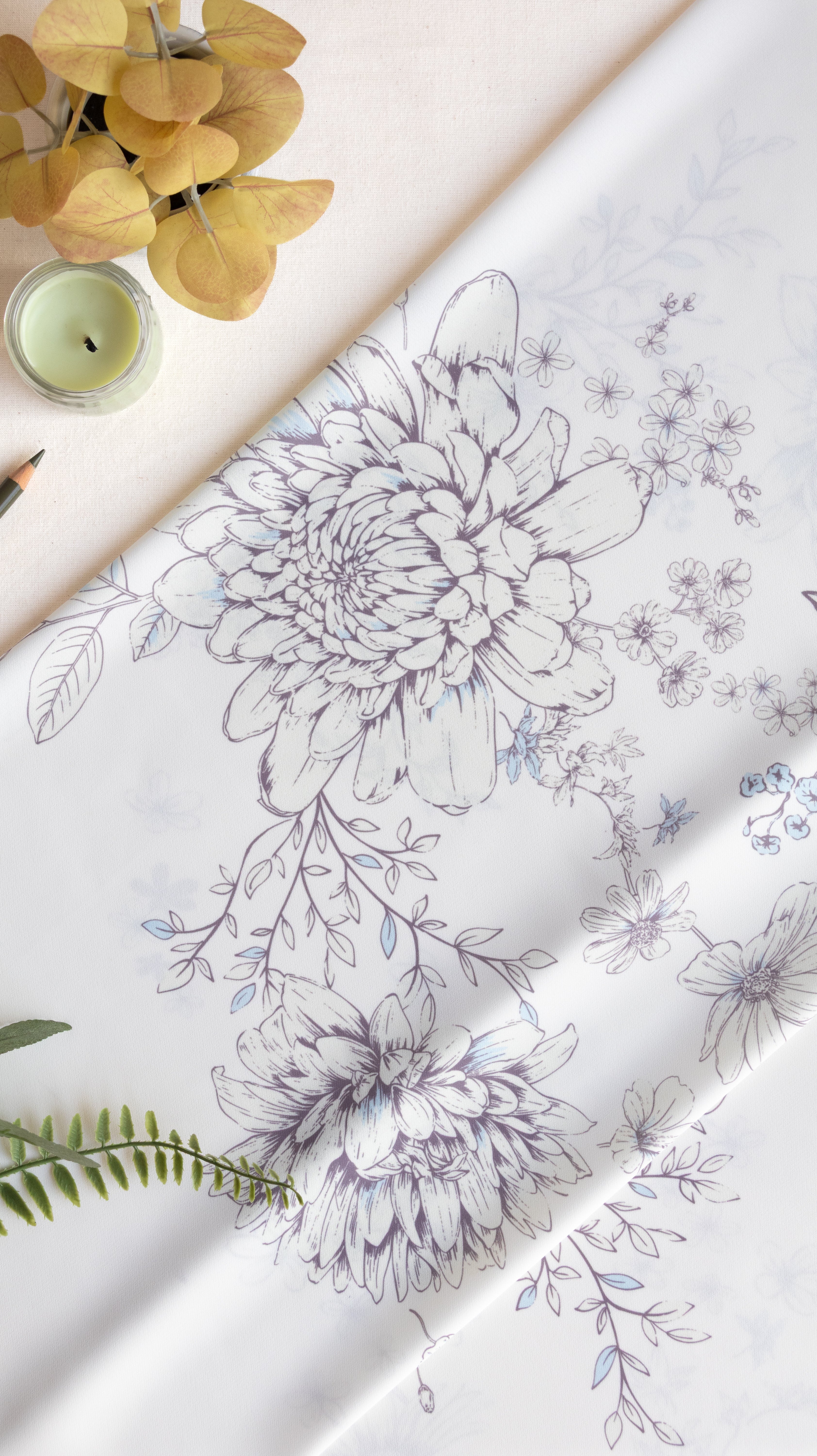White fabric with a delicate gray and blue floral line-art pattern, styled in a flat-lay composition with a candle, dried yellow flowers, and green leaves.