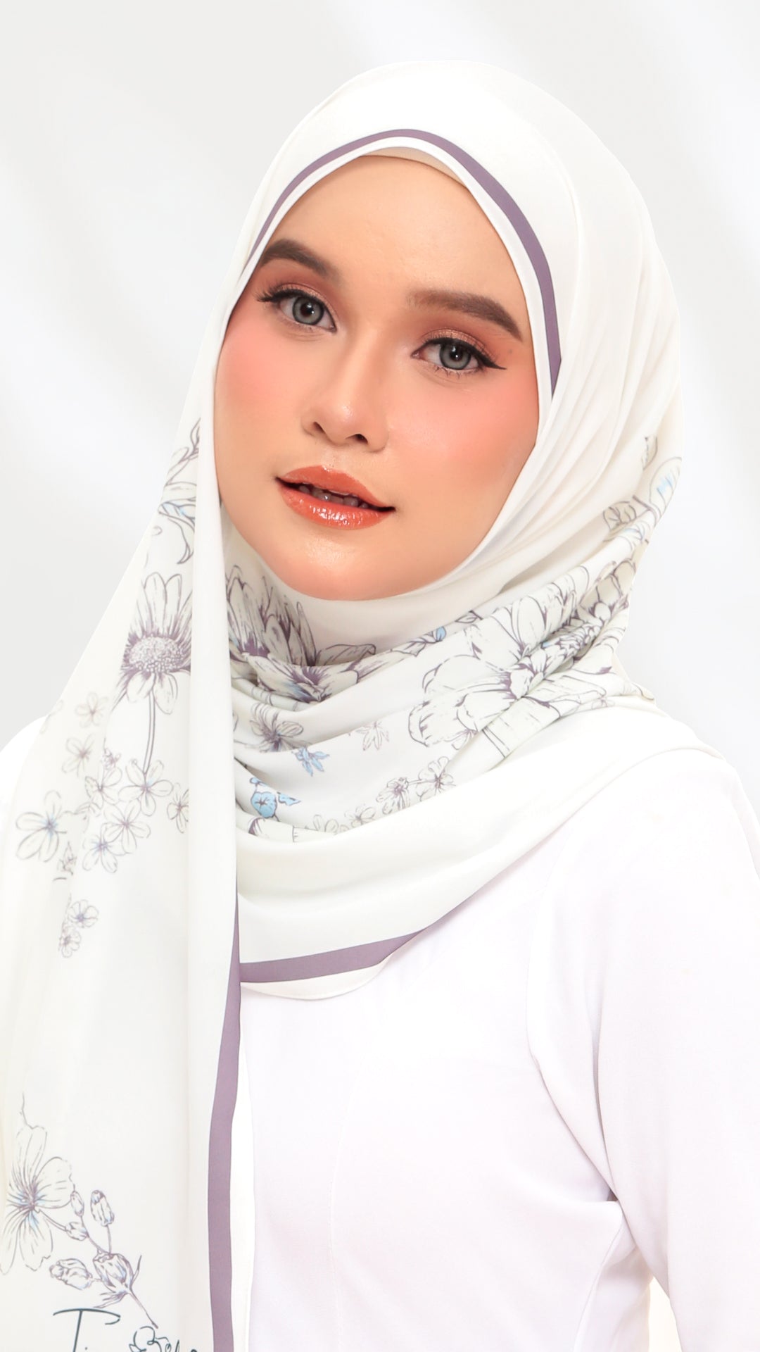 A woman wearing a white hijab with a delicate floral print and light purple trim, styled with a white top against a clean white background.