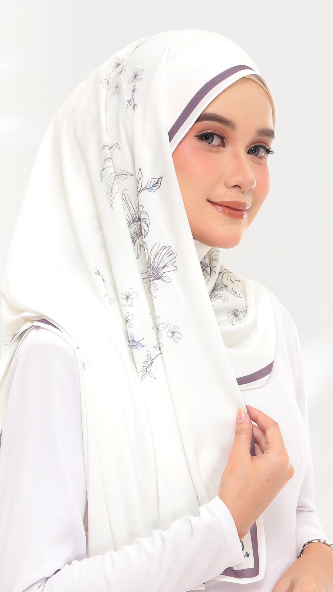 A woman wearing a white hijab with delicate gray floral patterns and thin purple trim, paired with a white long-sleeve top against a light gray studio background.