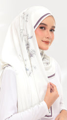A woman wearing a white hijab with delicate gray floral patterns and thin purple trim, paired with a white long-sleeve top against a light gray studio background.
