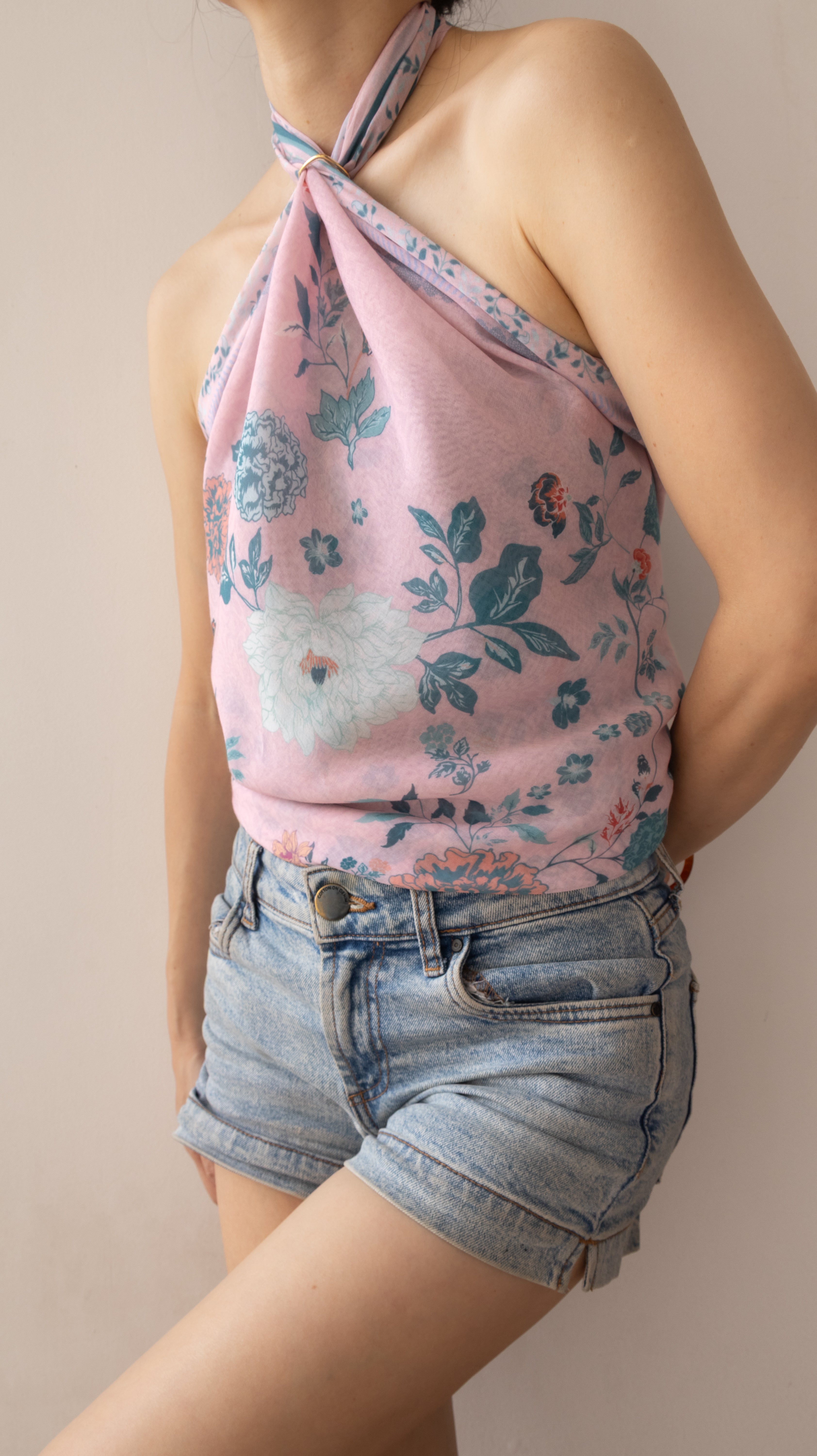 A woman wearing a light pink halter top with a blue and white floral print, paired with light-wash denim shorts, shown against a plain beige background.