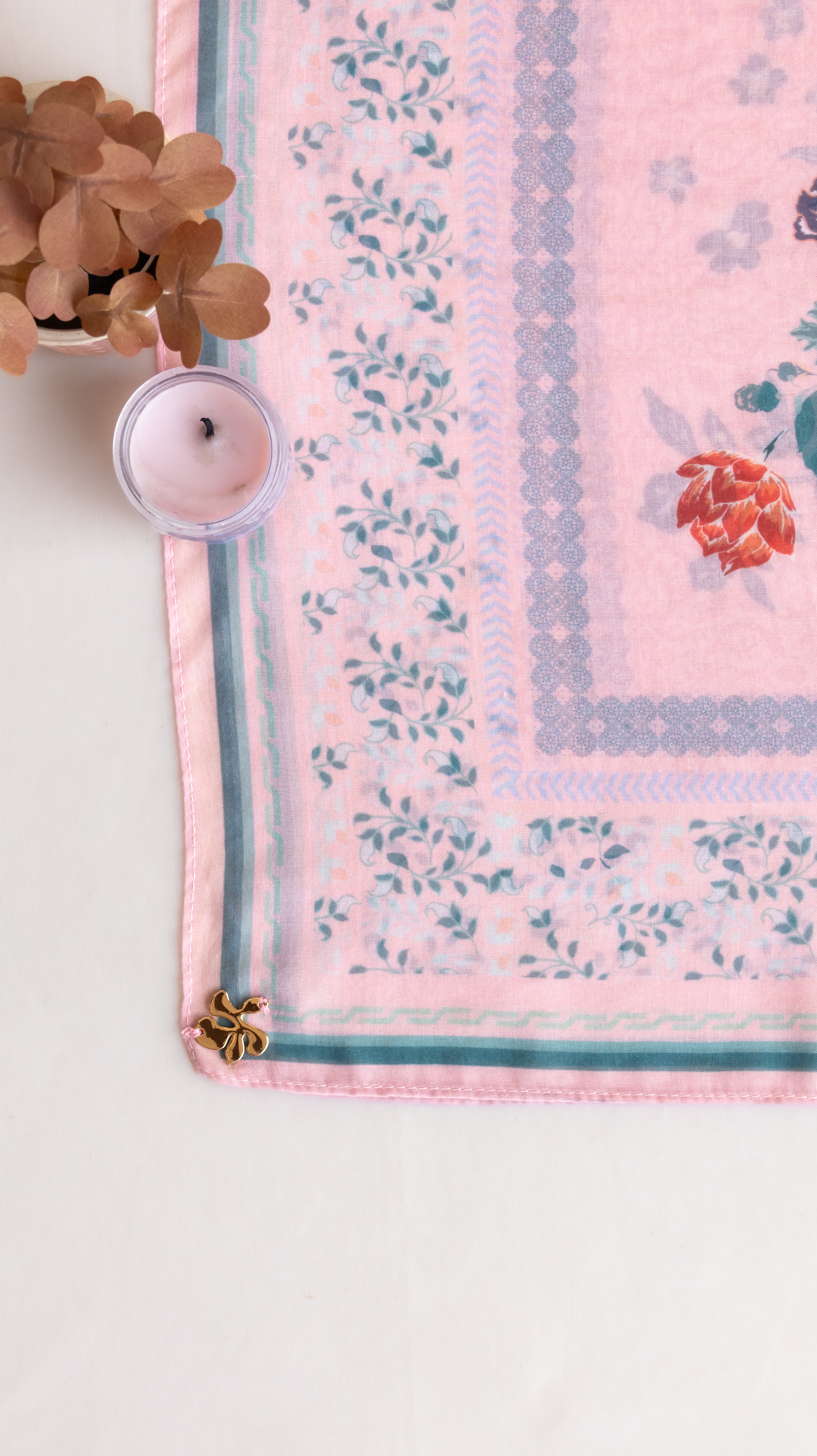 A pink patterned textile, likely a throw blanket or scarf, styled in a flat-lay with a small pink candle and dried flowers on a white surface.