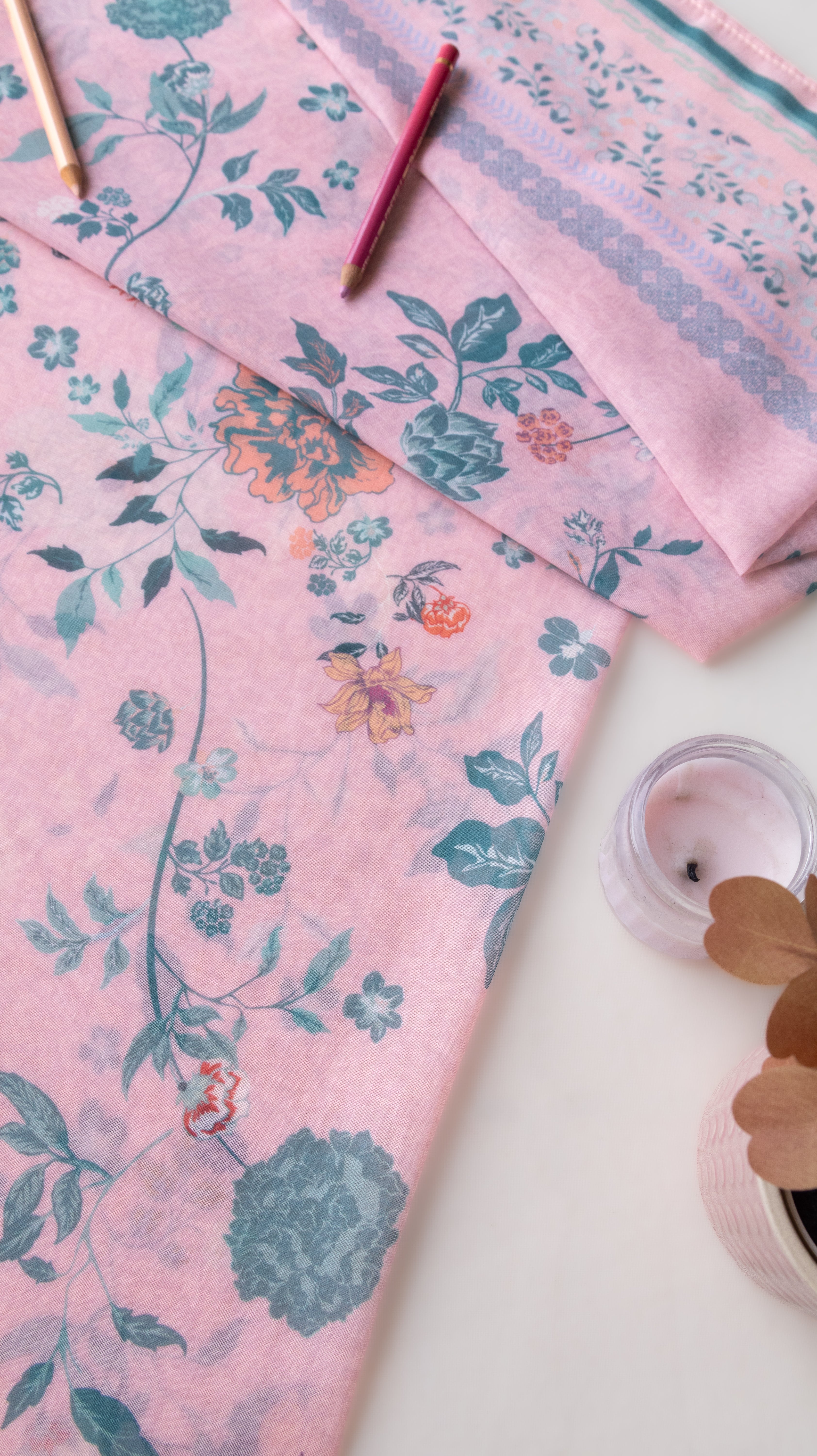 A folded pink fabric with a blue and orange floral vine pattern, styled on a white surface with a small candle and wooden pencils.