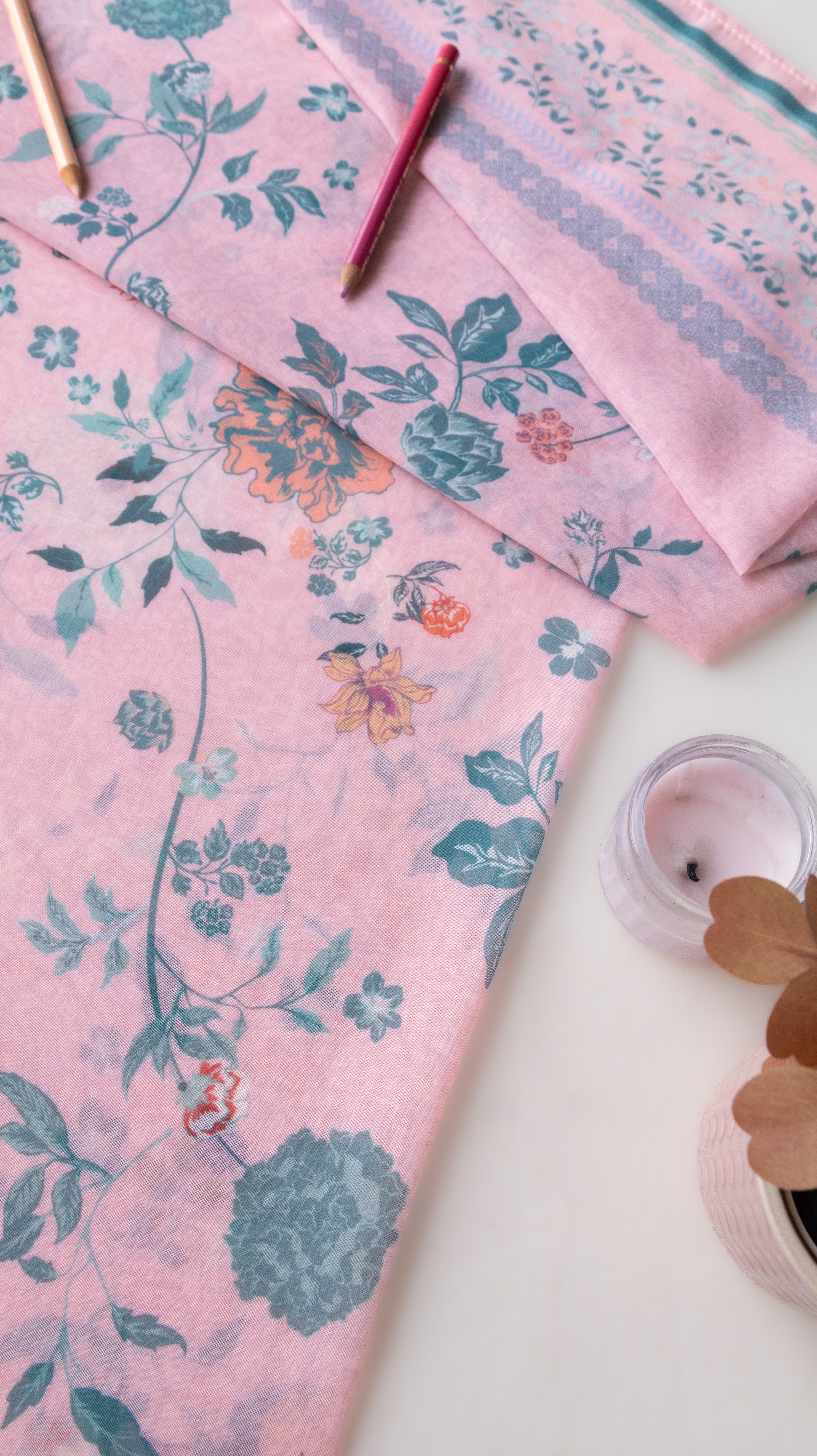 A folded pink fabric with a blue and orange floral vine pattern, styled on a white surface with a small candle and wooden pencils.
