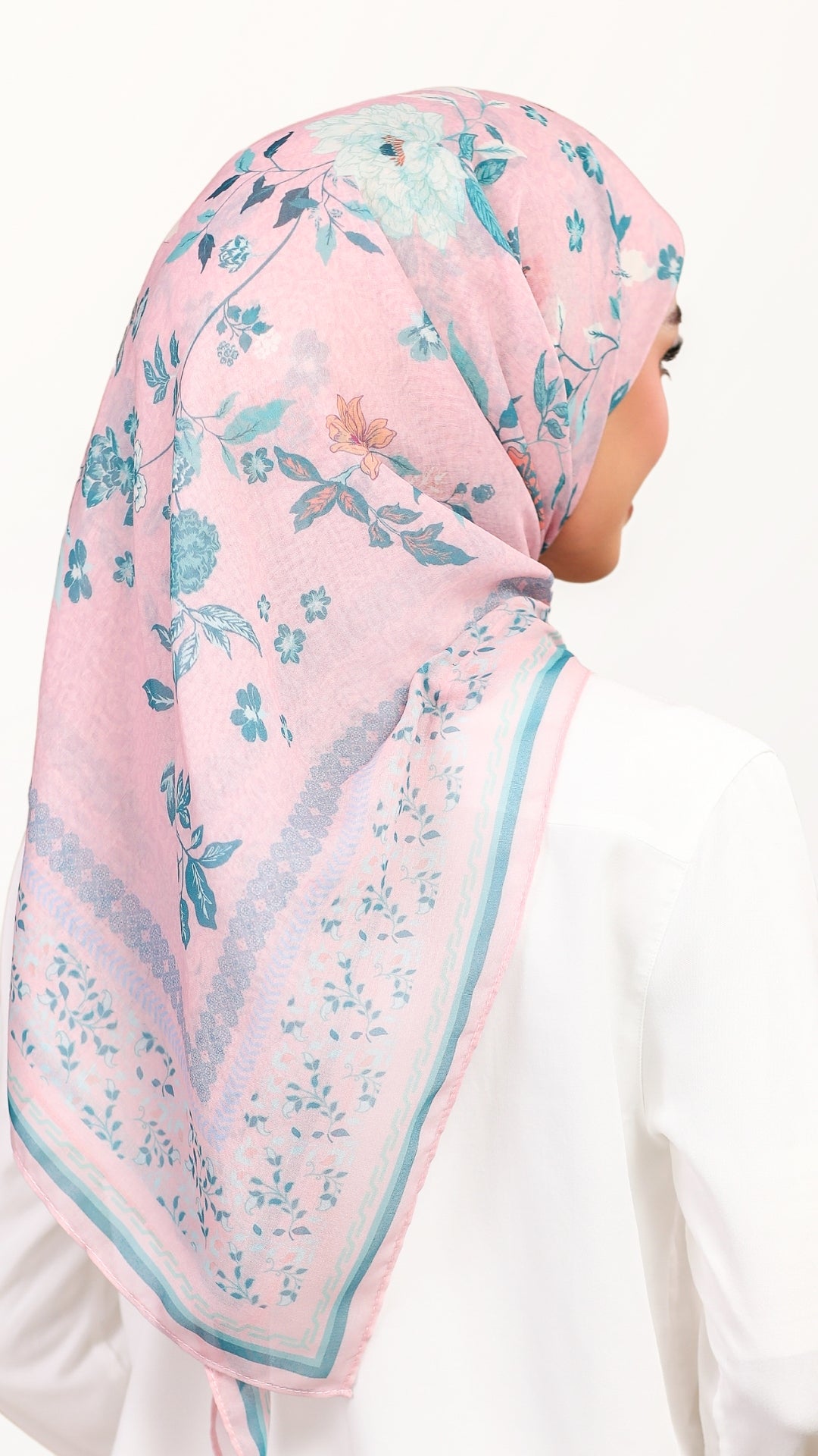 A woman shown from the back wearing a light pink hijab with a blue and green floral pattern and a matching border, paired with a white top against a plain white background.