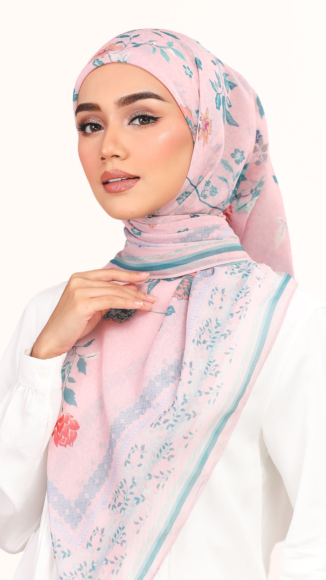 A woman wearing a light pink hijab with a blue floral pattern and a thin teal border, paired with a white top against a plain white background.